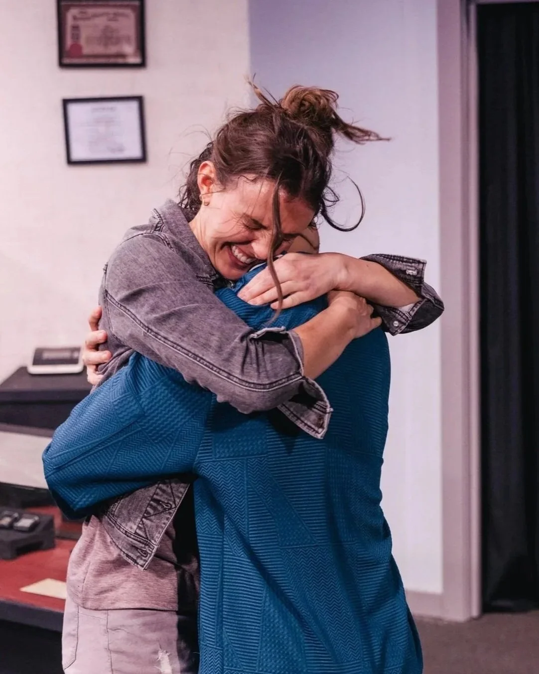 Two women hugging warmly with big smiles, embracing each other tightly in an indoor setting.