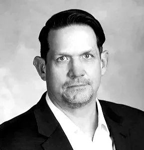 Black and white portrait of a man with dark hair, wearing a suit and a white shirt, looking directly at the camera.