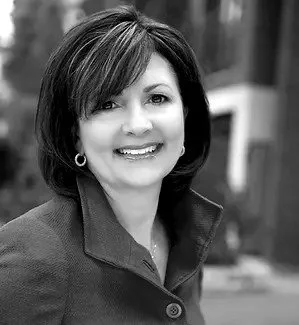 Headshot of a smiling woman with short dark hair, wearing a collared jacket, standing outdoors in an urban setting.