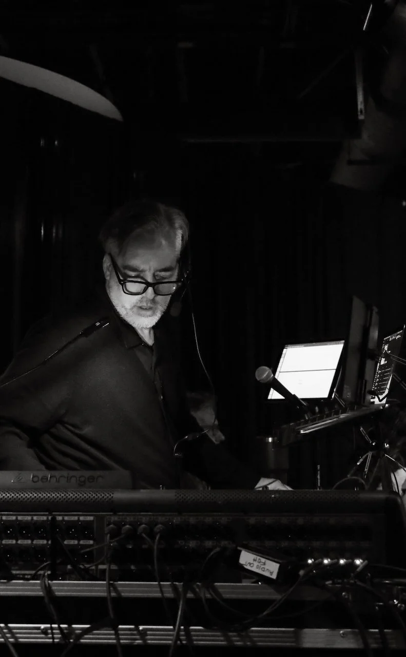 A man with glasses and a beard wearing headphones is operating music equipment in a dimly lit setting, possibly a stage or recording studio.