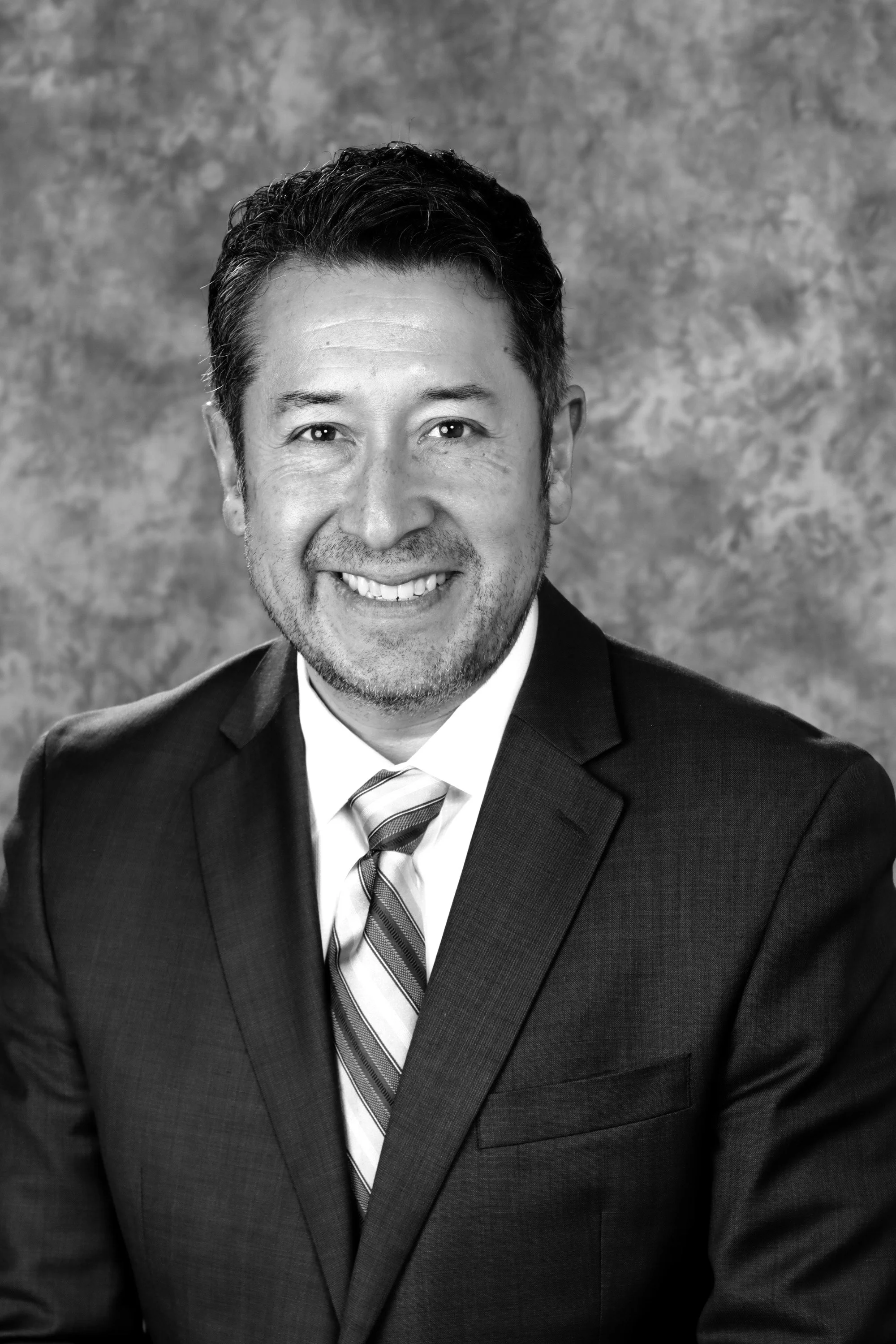A black and white professional headshot of a smiling man wearing a dark suit, white shirt, and striped tie, against a textured background.
