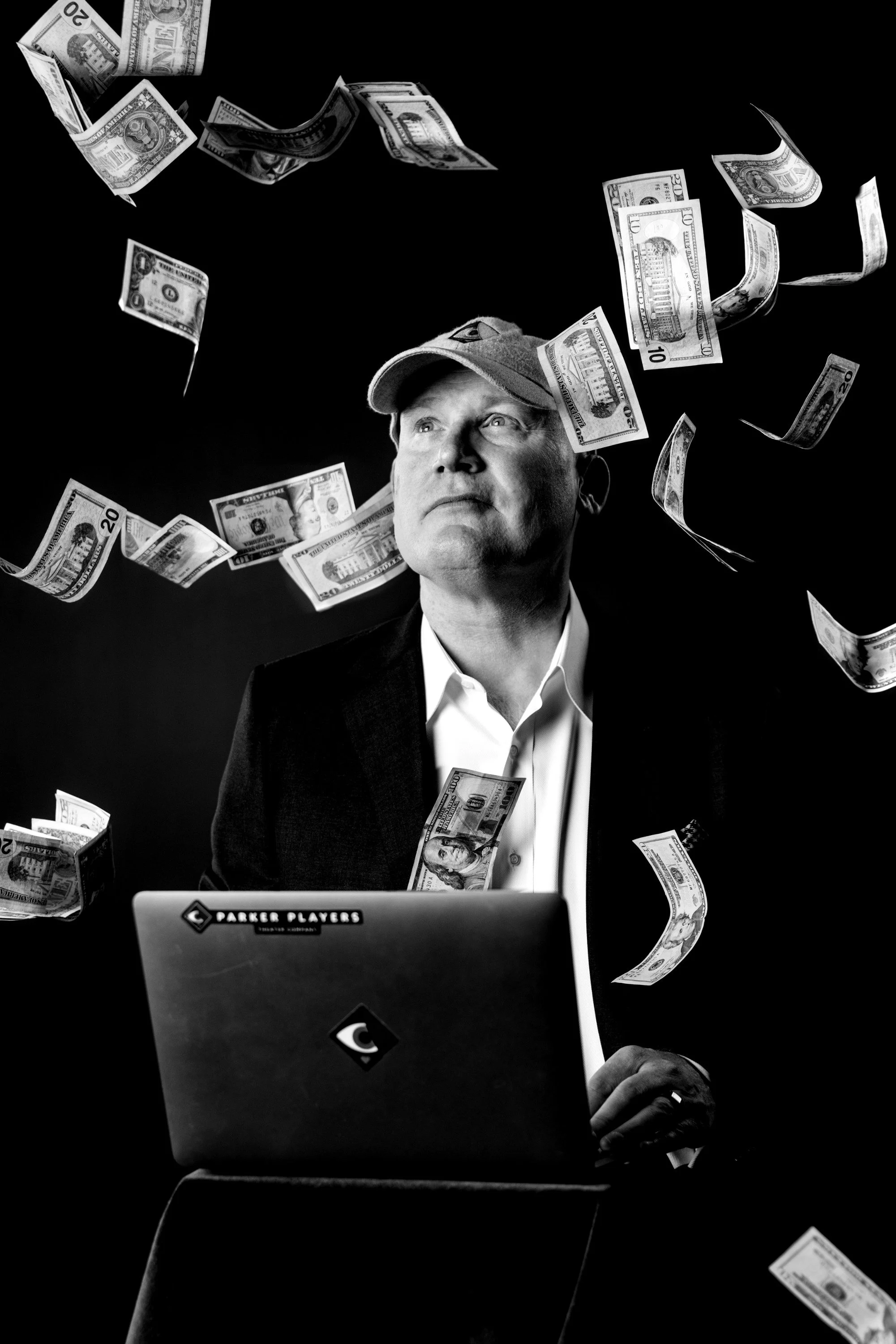 A man with a cap sits at a desk with a laptop while dollar bills are falling around him in a black and white photo.