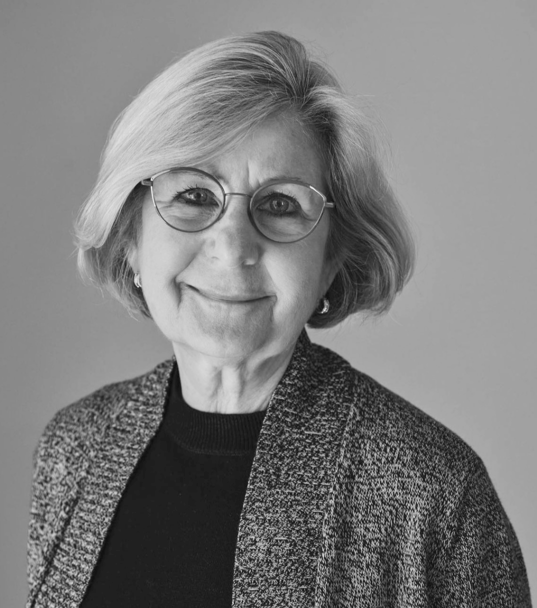 Black and white portrait of a smiling older woman with glasses, short wavy hair, wearing a dark top and a textured blazer.
