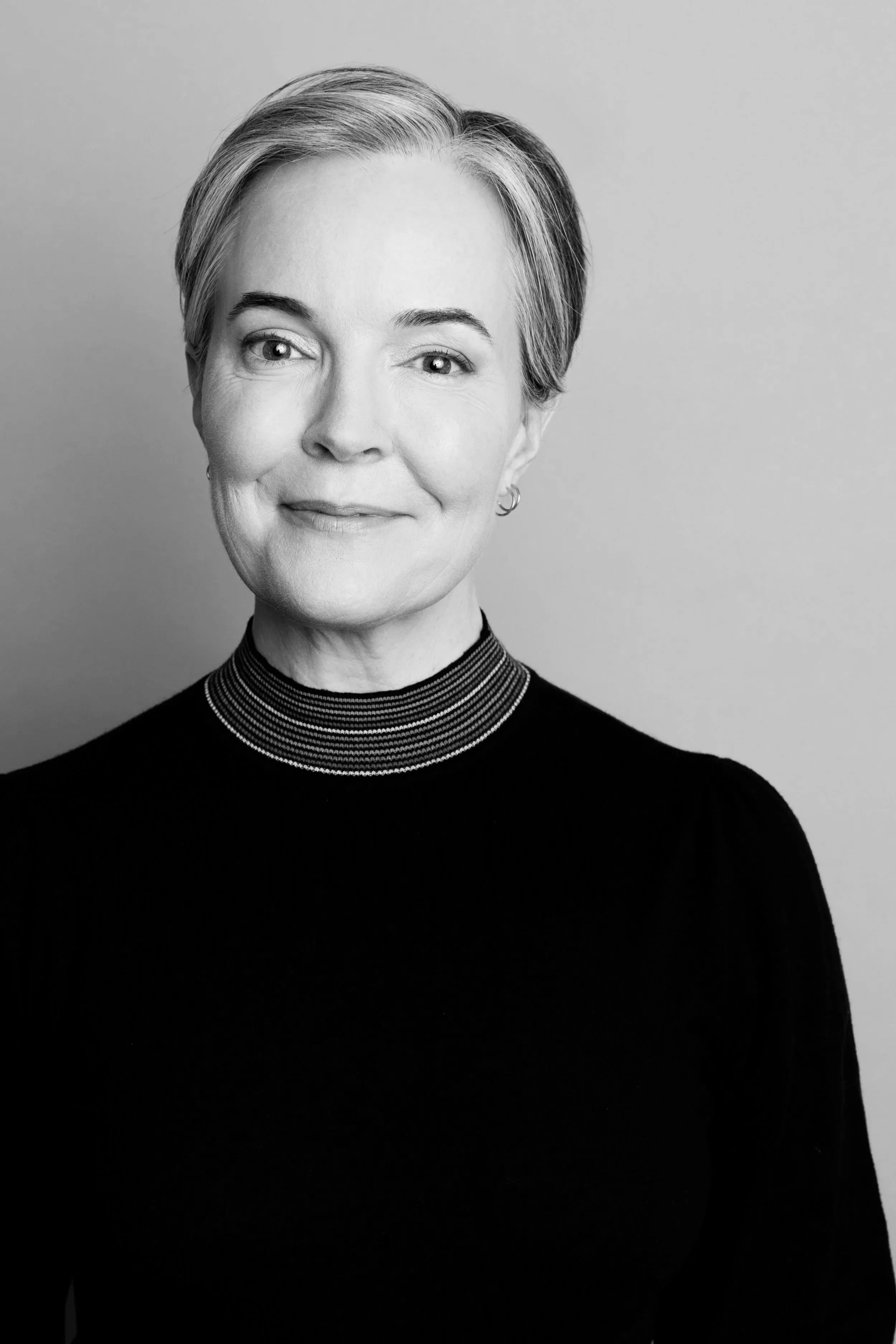 Black-and-white portrait of a woman with short, light-colored hair, wearing a high-necked black top with a patterned collar, smiling slightly.
