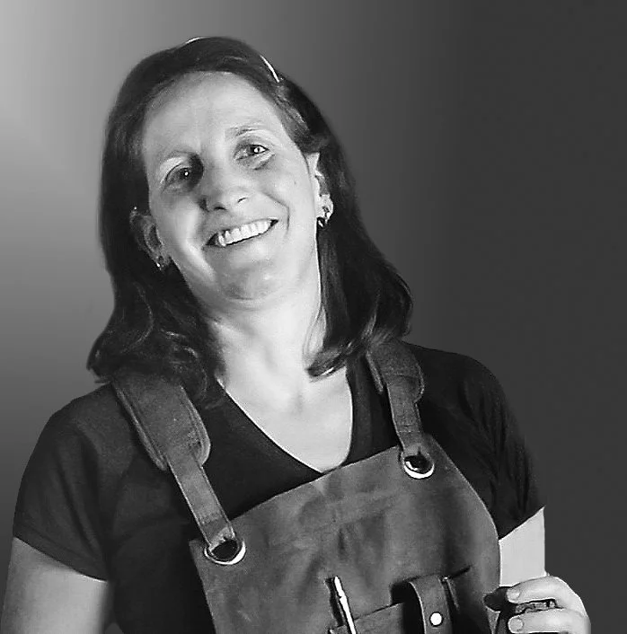 A woman with dark hair smiling, wearing a dark t-shirt and an apron, against a plain background.