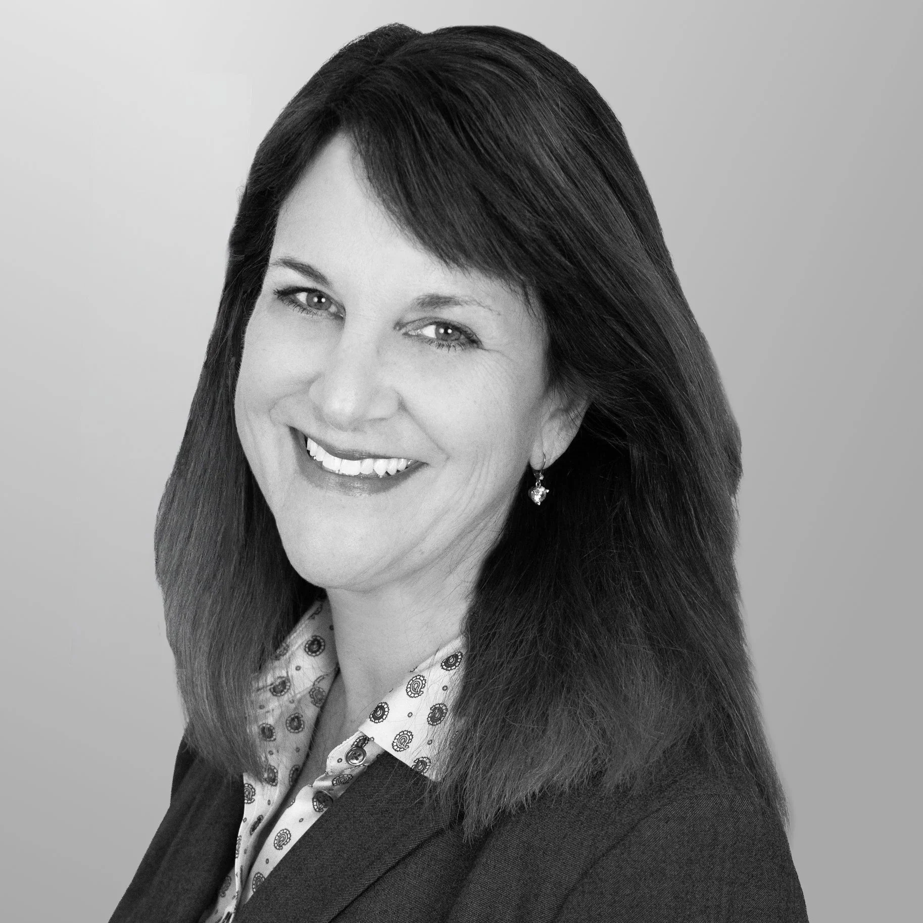 Black-and-white professional headshot of a woman with shoulder-length dark hair, smiling, wearing earrings, a patterned blouse, and a blazer.