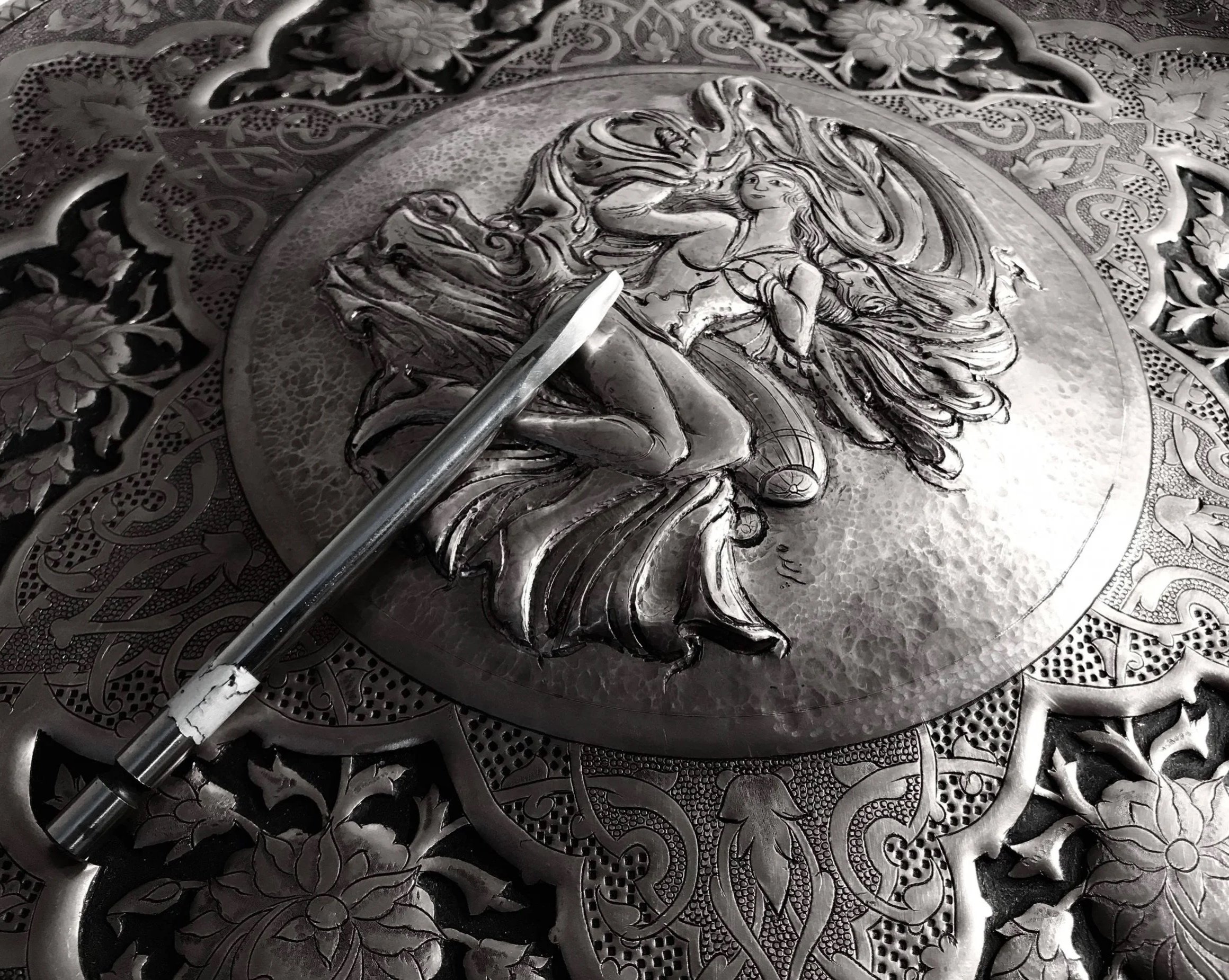 Intricate metalwork featuring a raised depiction of a woman with flowing hair, surrounded by detailed floral and geometric patterns.