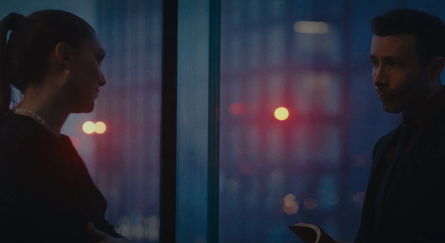 A man and woman are standing on opposite sides of a glass window during dusk, with city lights blurred in the background, engaging in a conversation.