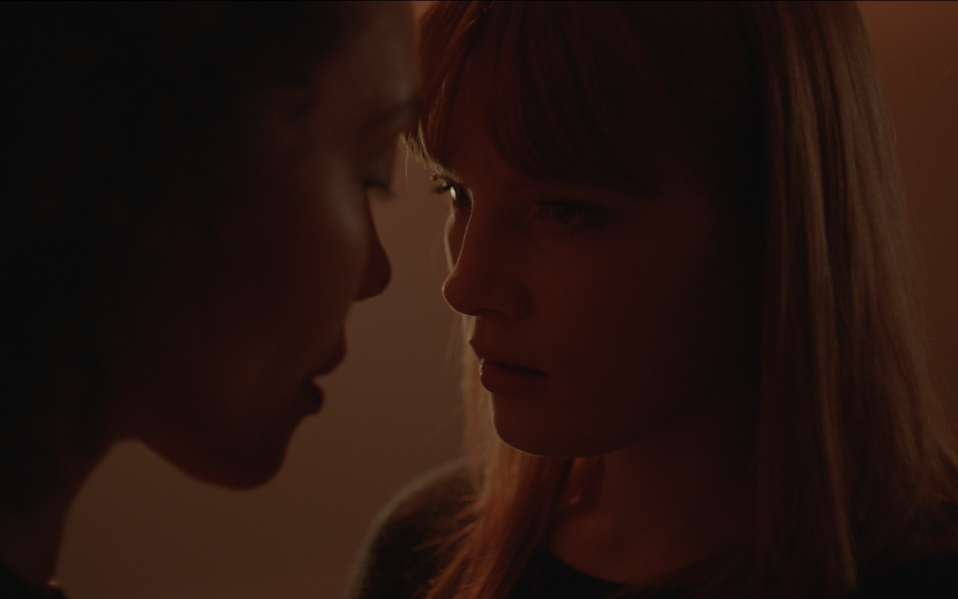 Two women with close-up profiles touching foreheads, dimly lit, with focused expressions.