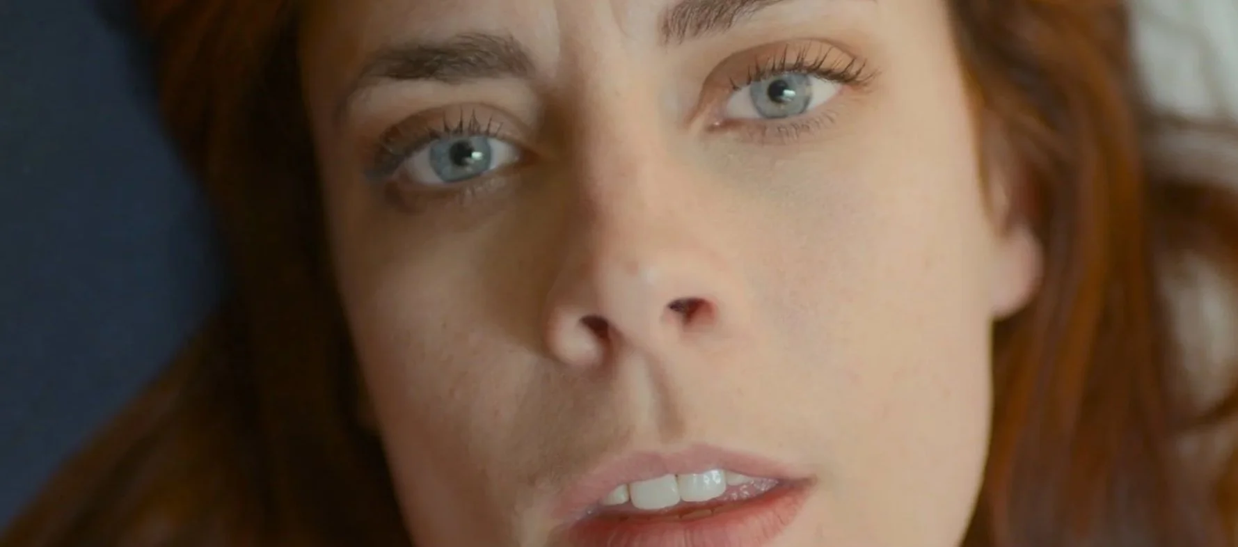 Close-up of a woman with light skin and blue eyes lying down, gazing upwards with a contemplative expression.