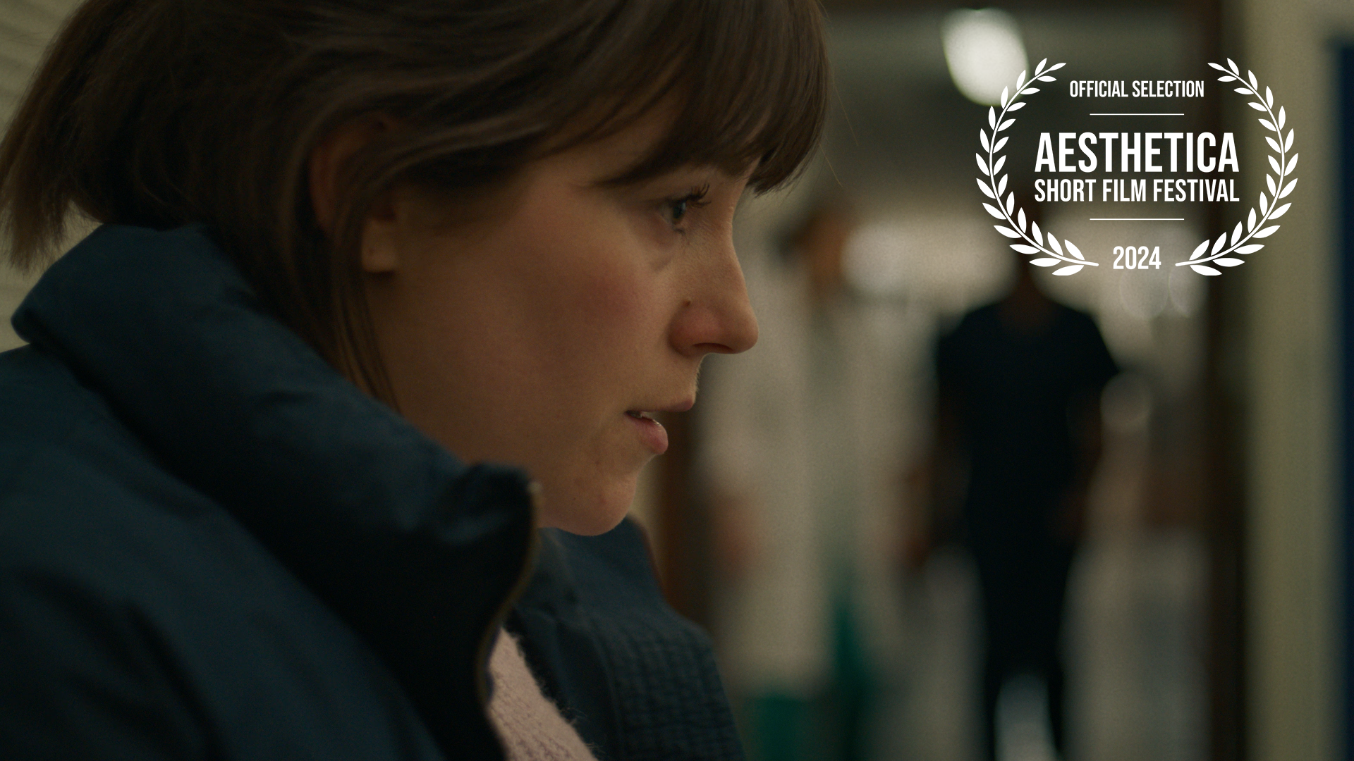 Close-up of a woman in profile with short brown hair, wearing a dark jacket, standing in a school hallway with blurred background, and an overlay badge indicating recognition at the 2024 Aesthetica Short Film Festival.