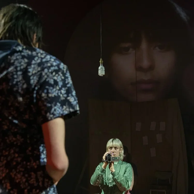 A woman with blonde hair dressed in green, holding a camera, facing a large mural of a person's face on the wall. A person with dark hair and a floral shirt is seen from behind on the left side of the image.
