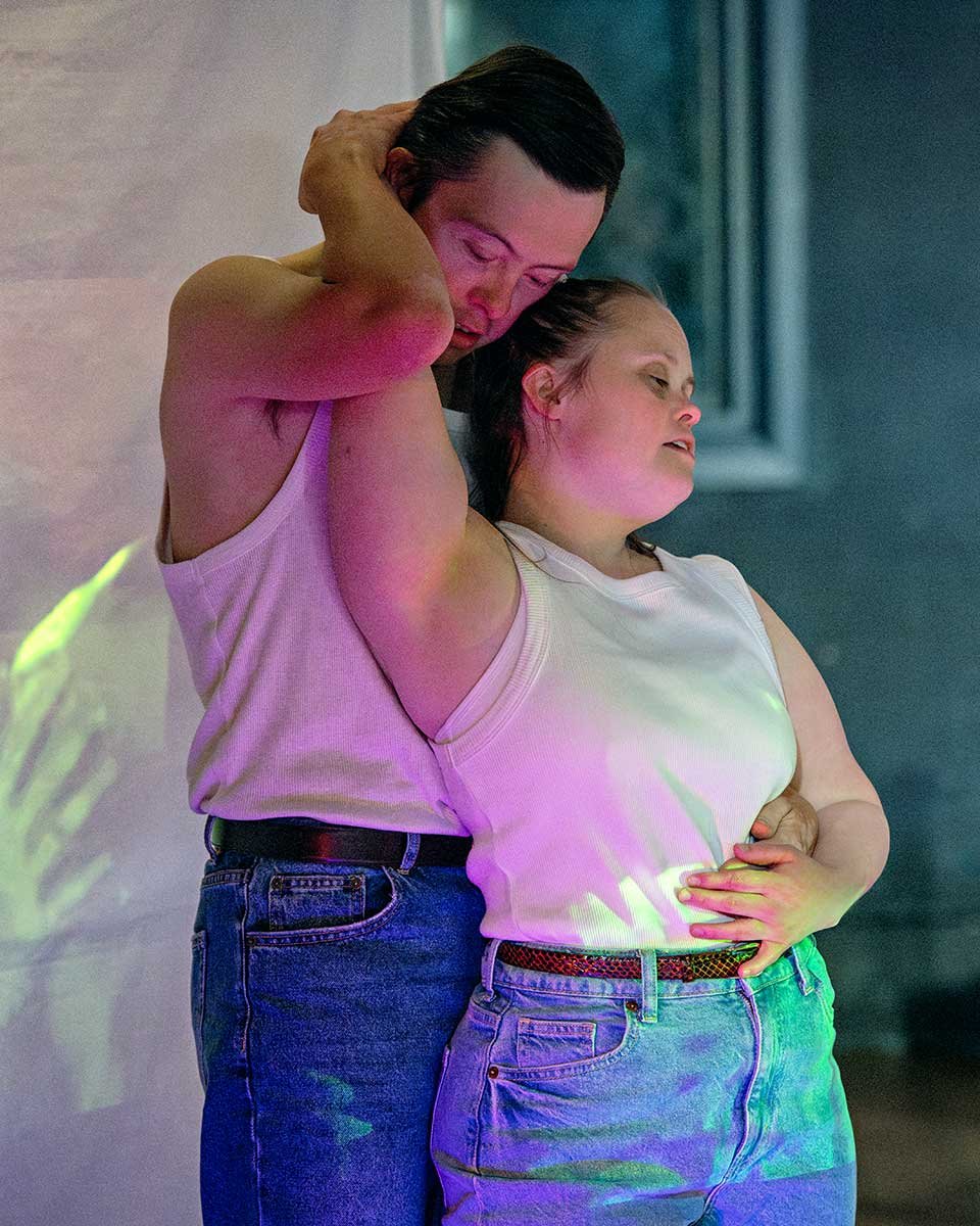 A man and a woman are embracing in a close, intimate pose with colorful lighting.