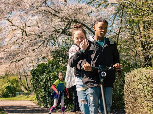 Two kids riding a scooter and a girl walking on a park trail surrounded by trees and blooming flowers.