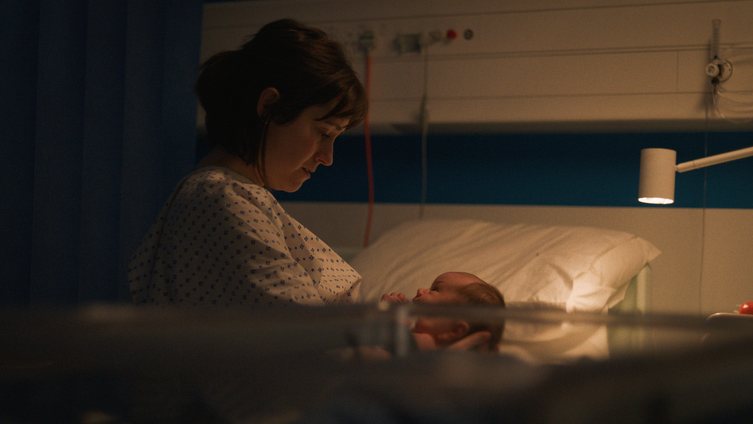 A woman in a hospital gown looking down at a newborn baby in an incubator.