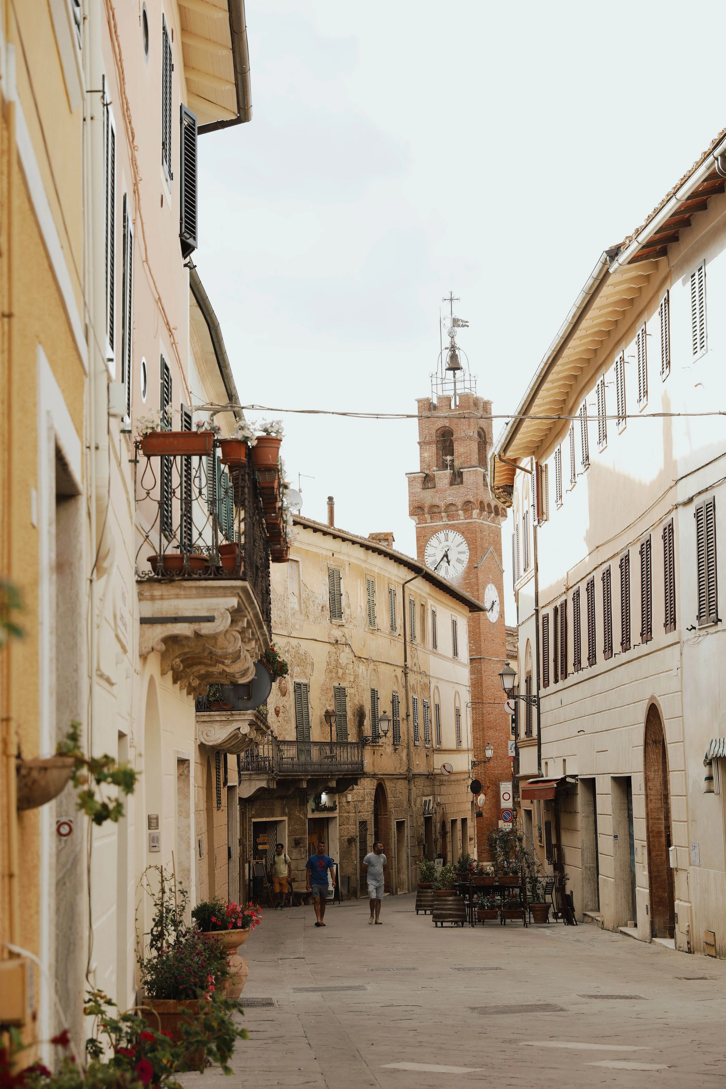 A narrow European street with old buildings, flower pots, and a clock tower in the background.