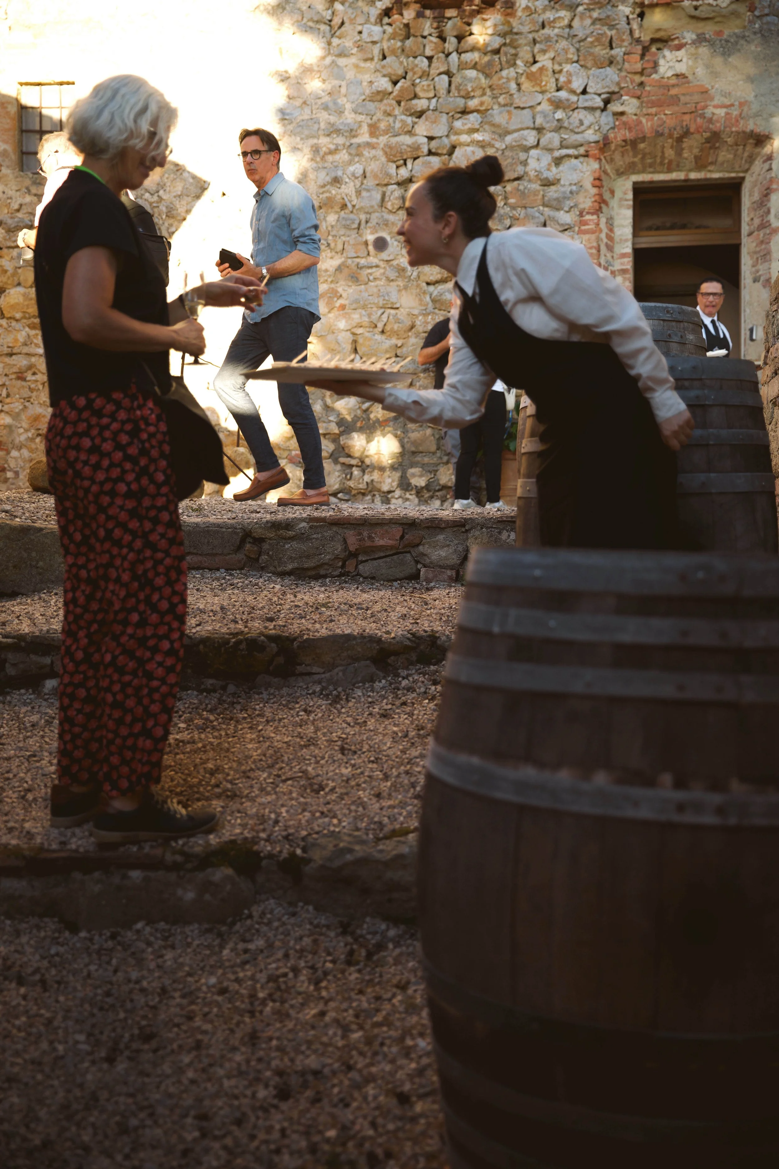 A woman in a black top with red patterned pants converses with a waitress holding a tray at an outdoor event, with a stone building and other people in the background.