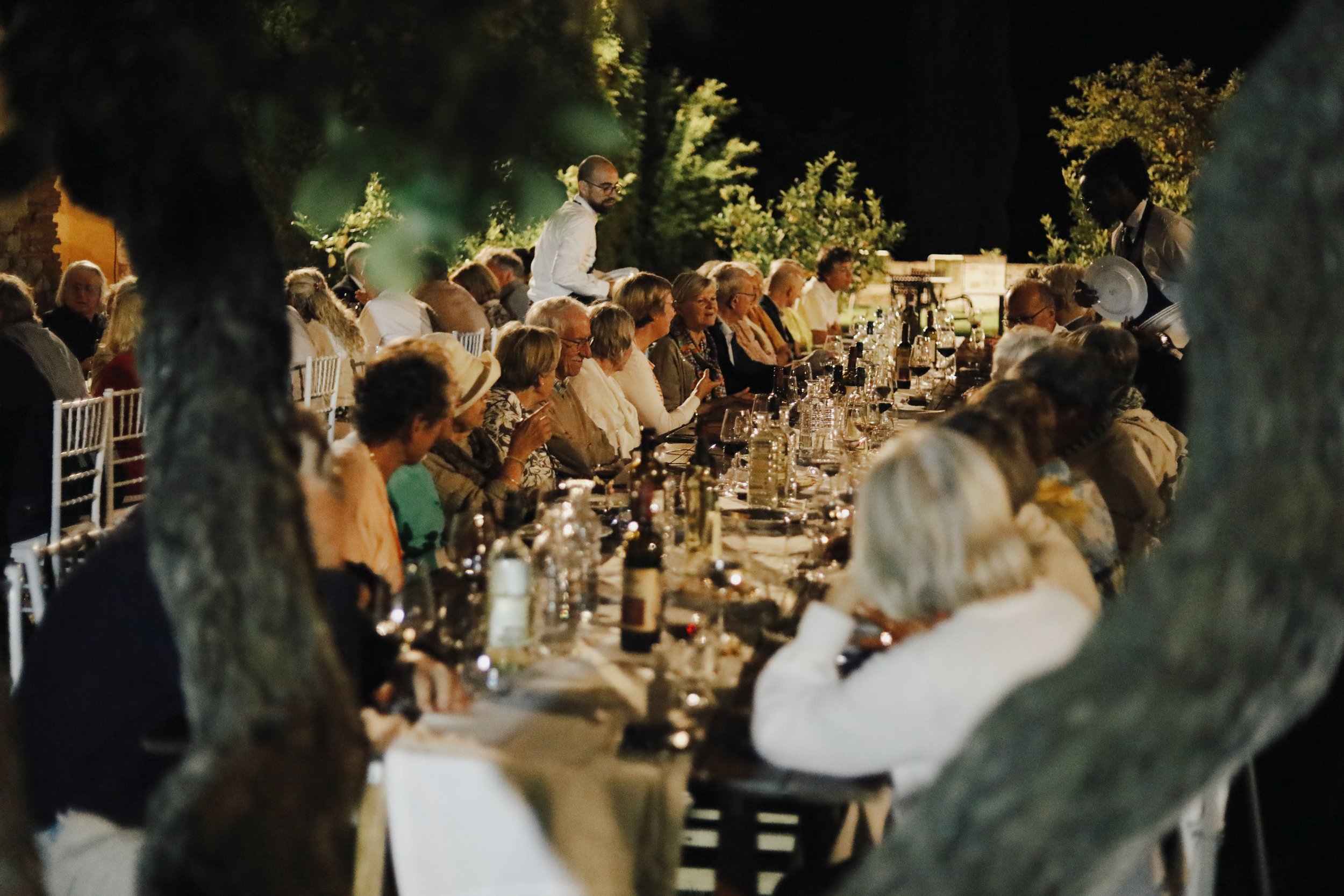 People seated at a long outdoor dinner table at night, with trees and warm lighting in the background, some waitstaff serving drinks.