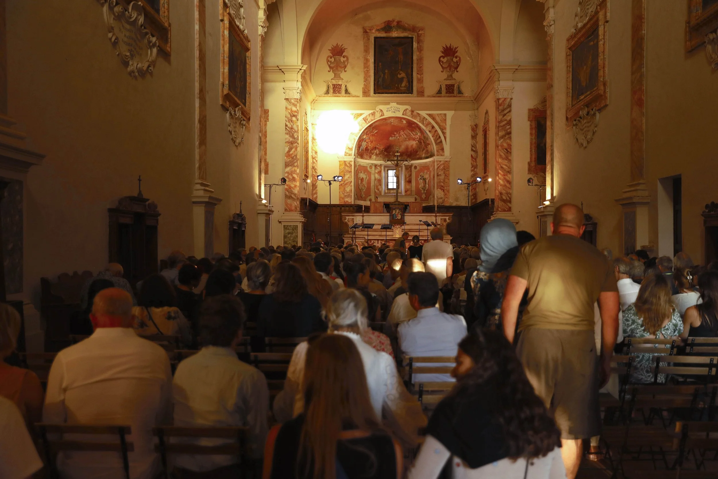 People attending a concert or event inside a church with ornate interior, high ceilings, and religious artwork, facing the altar at the front.