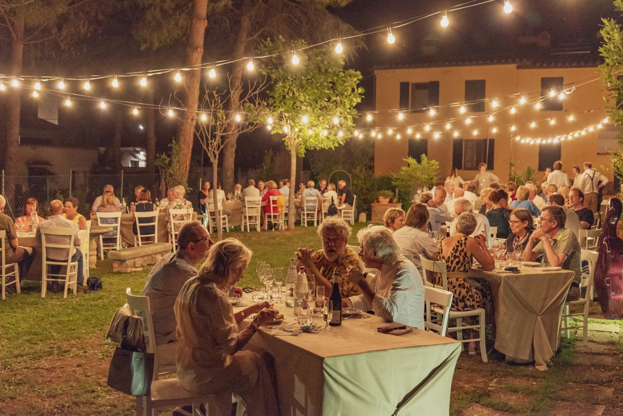 People dining at outdoor tables under string lights at night, in a garden setting.