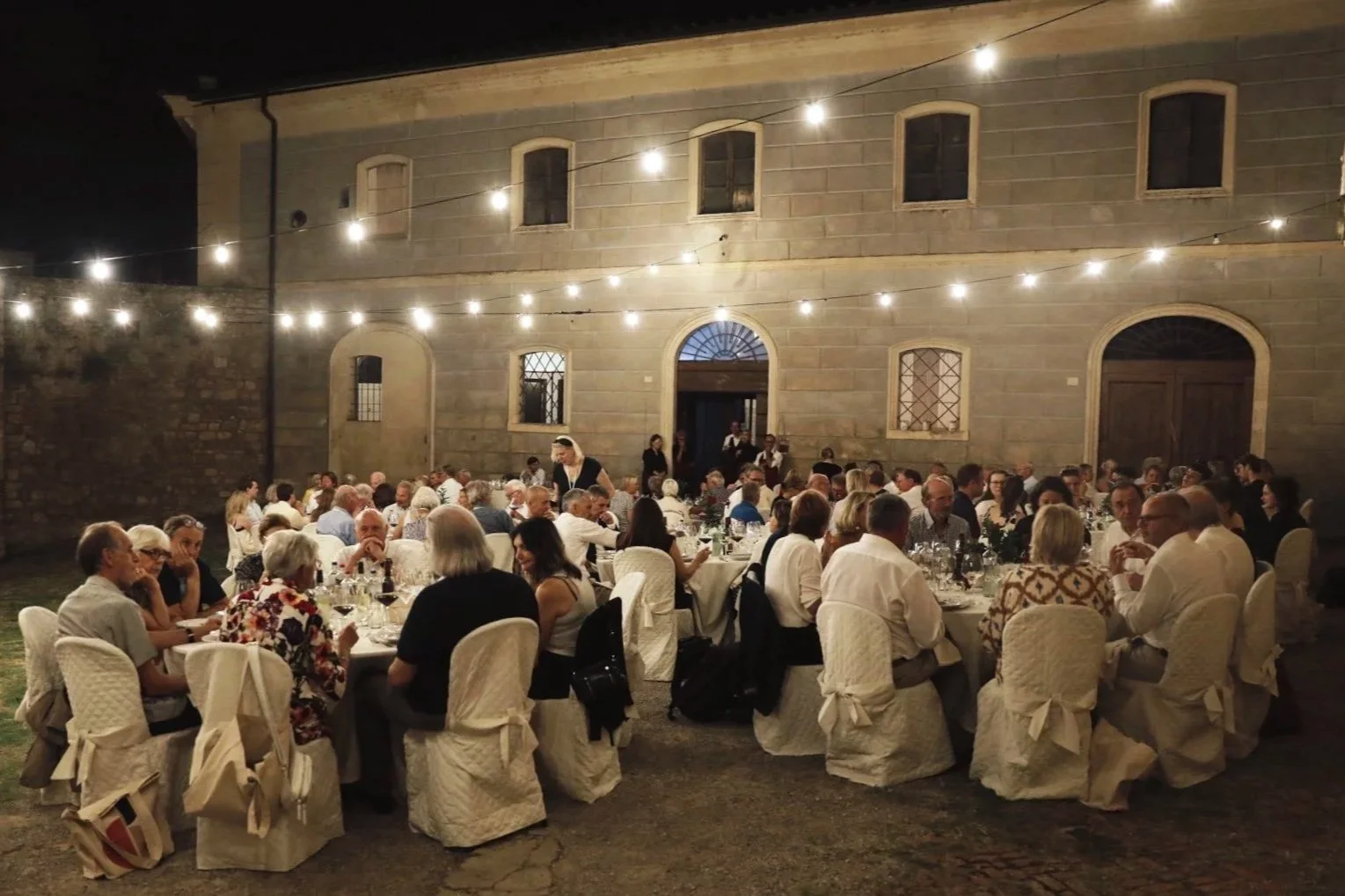 An outdoor evening banquet with numerous guests seated at round tables, decorated with tableware and wine glasses, under string lights in front of a historic stone building.