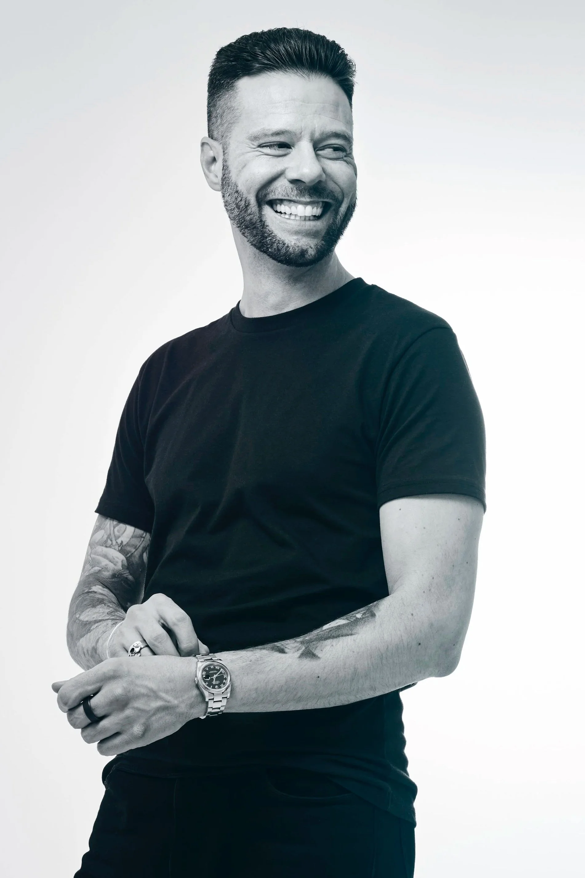 A smiling man with short dark hair and a beard, wearing a black t-shirt, a watch, and rings, standing against a gradient background.