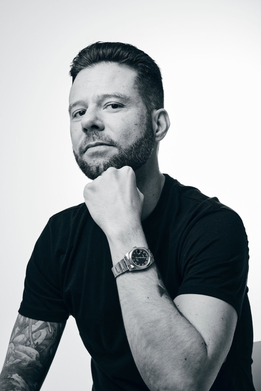 Black and white portrait of a man with a beard, tattooed arm, wearing a watch on his left wrist, resting his chin on his hand, looking serious.