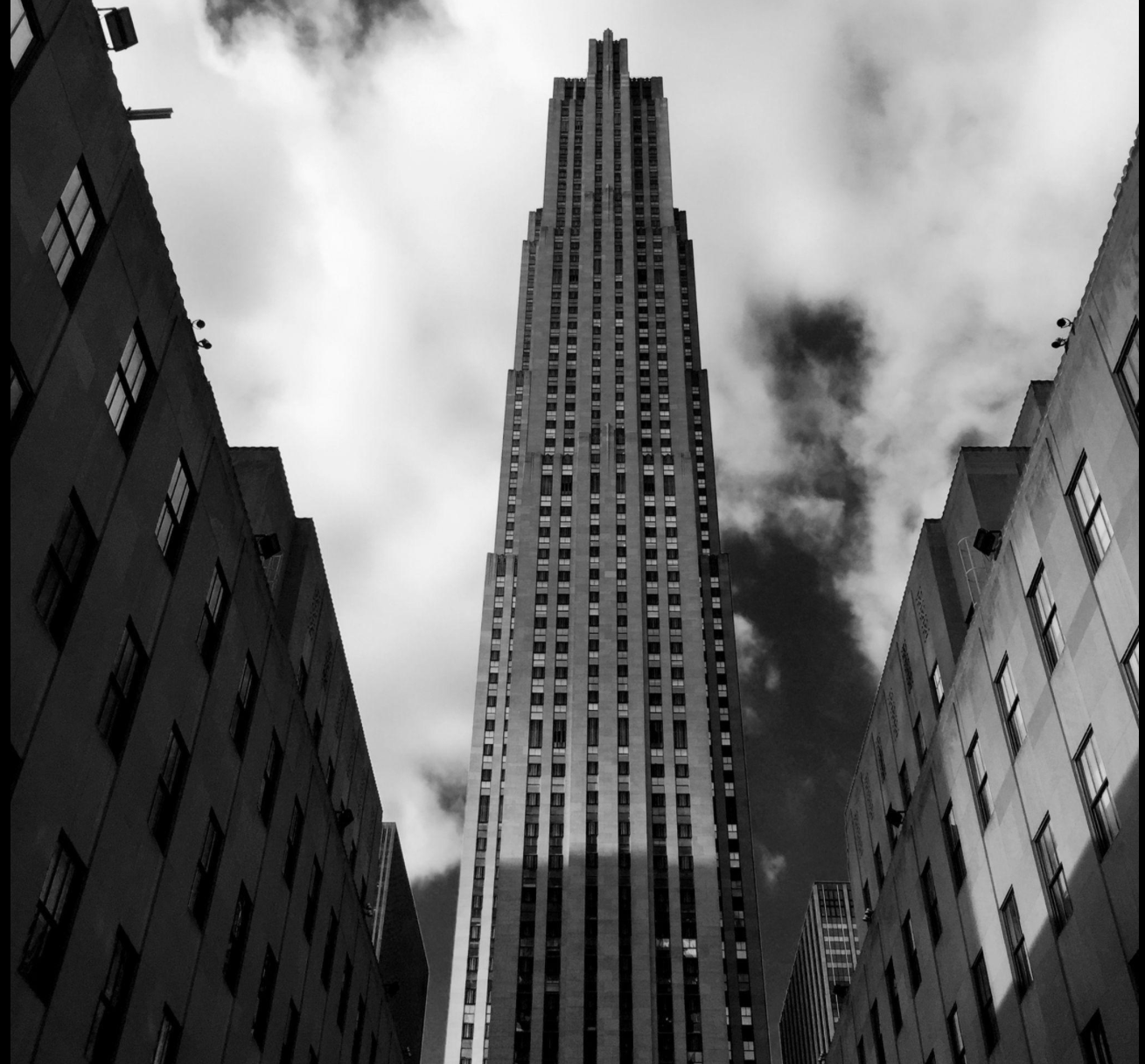 Black and white photo of a tall skyscraper viewed from ground level, with surrounding buildings and cloudy sky.