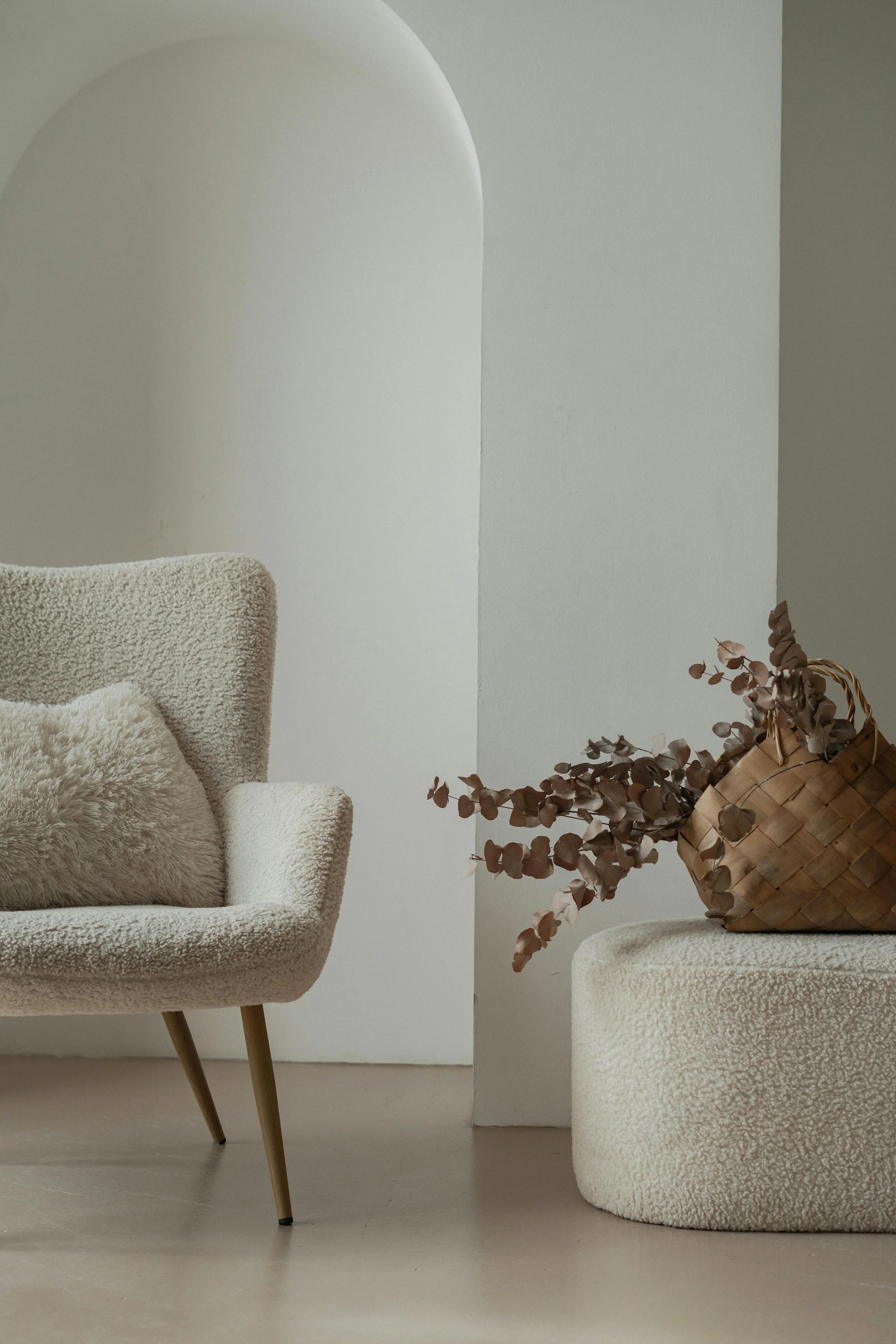 Un fauteuil beige avec un coussin en peluche, un panier en osier avec des branches de feuillage suspendues, dans une pièce au décor minimaliste.