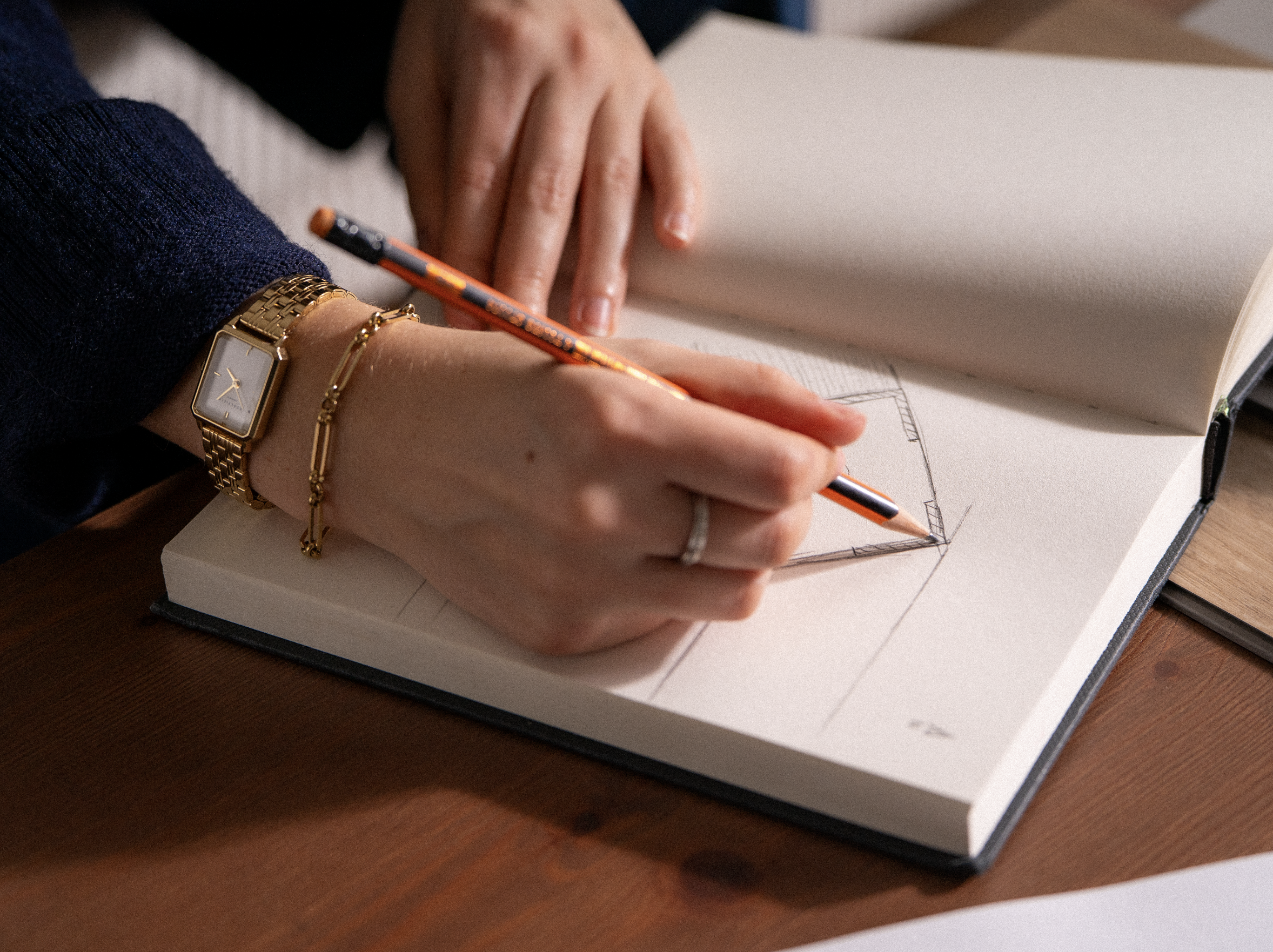 Une main tenant un crayon dessinant un croquis dans un carnet ouvert, avec une autre main posée sur le carnet. La personne porte une montre en or, deux bracelets en or, et une alliance.