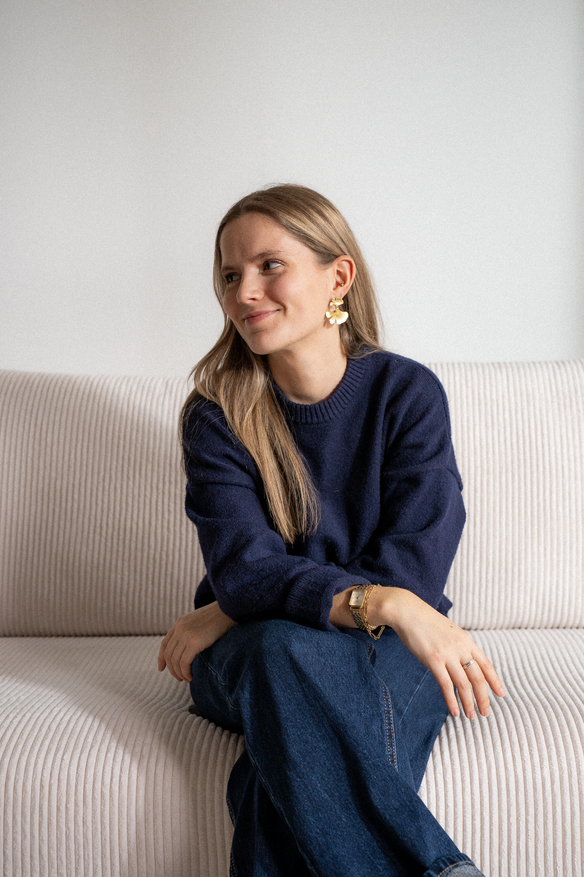 Jeune femme assise sur un canapé beige, portant un pull bleu foncé, des bijoux en or et des jeans, avec une expression souriante.