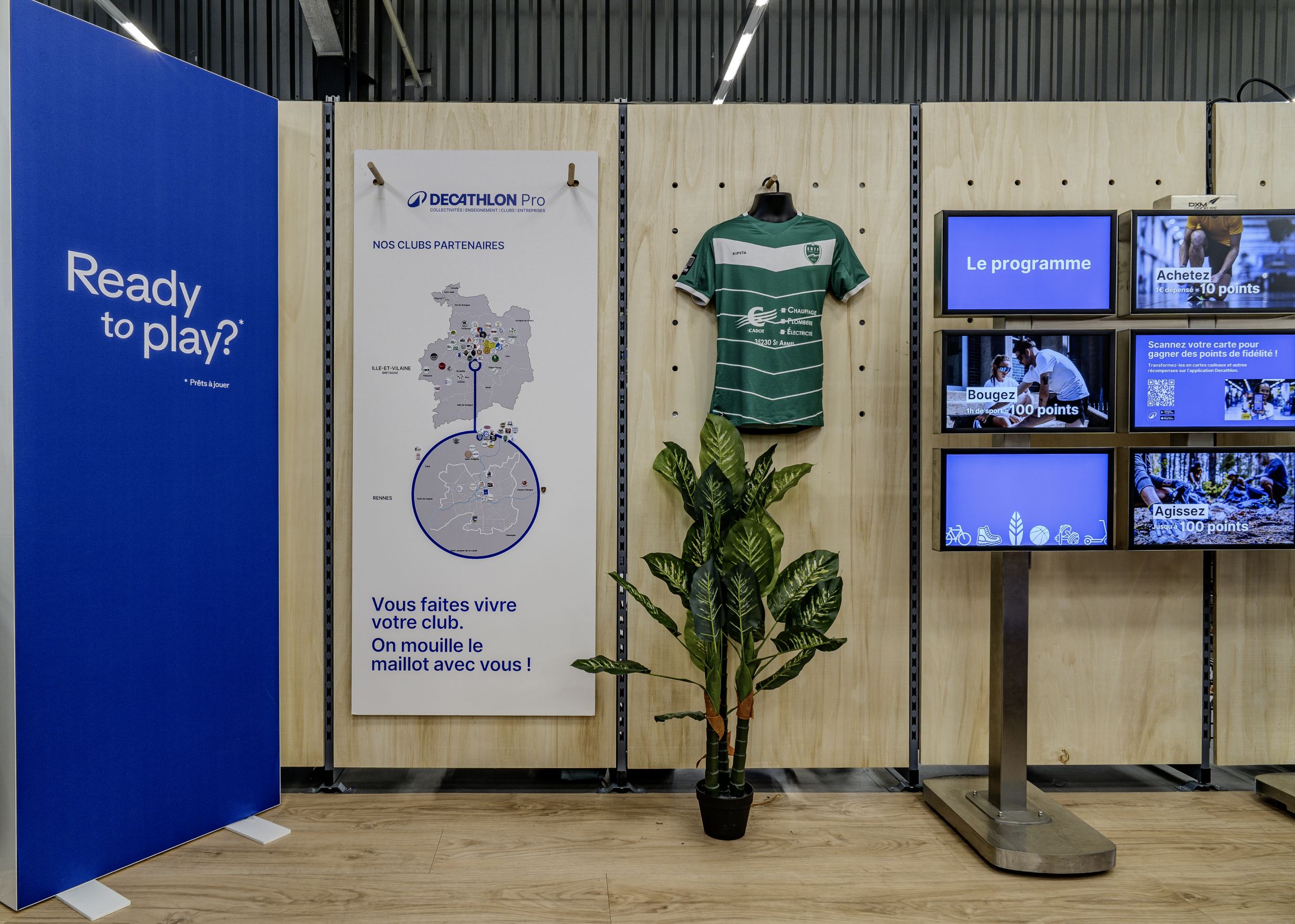 Espace d'accueil de Decathlon d'une salle avec un panneau d'affichage, un maillot de football vert, une plante verte, et plusieurs écrans affichant le programme de fidélité Decathlon. Par Alexane Azé Architecture.
