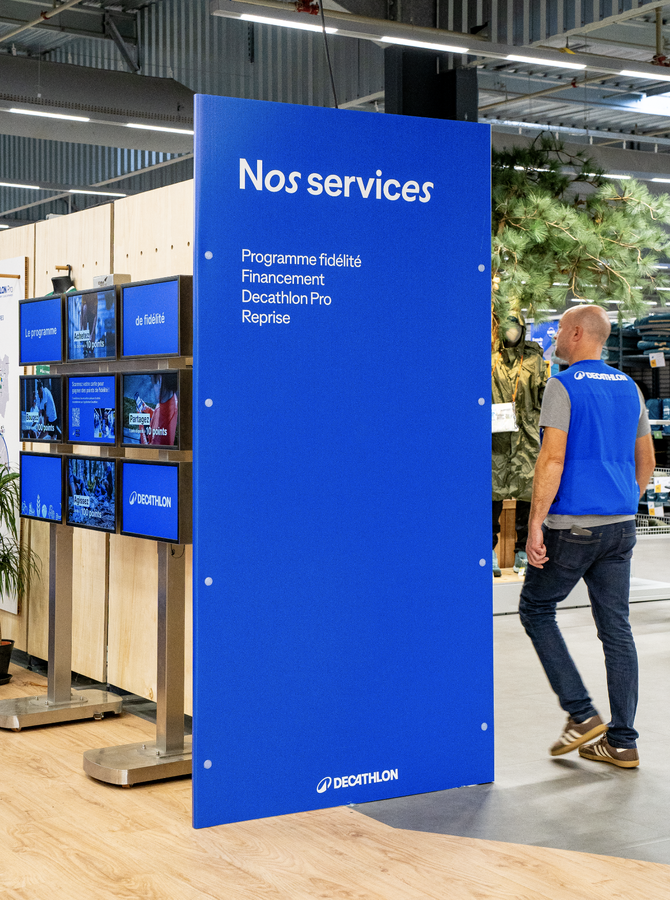 Panneau bleu avec la légende 'Nos services' devant un espace Decathlon avec un employé en gilet bleu, dans un magasin Decathlon, exposant des produits de sport. Par Alexane Azé Architecture.