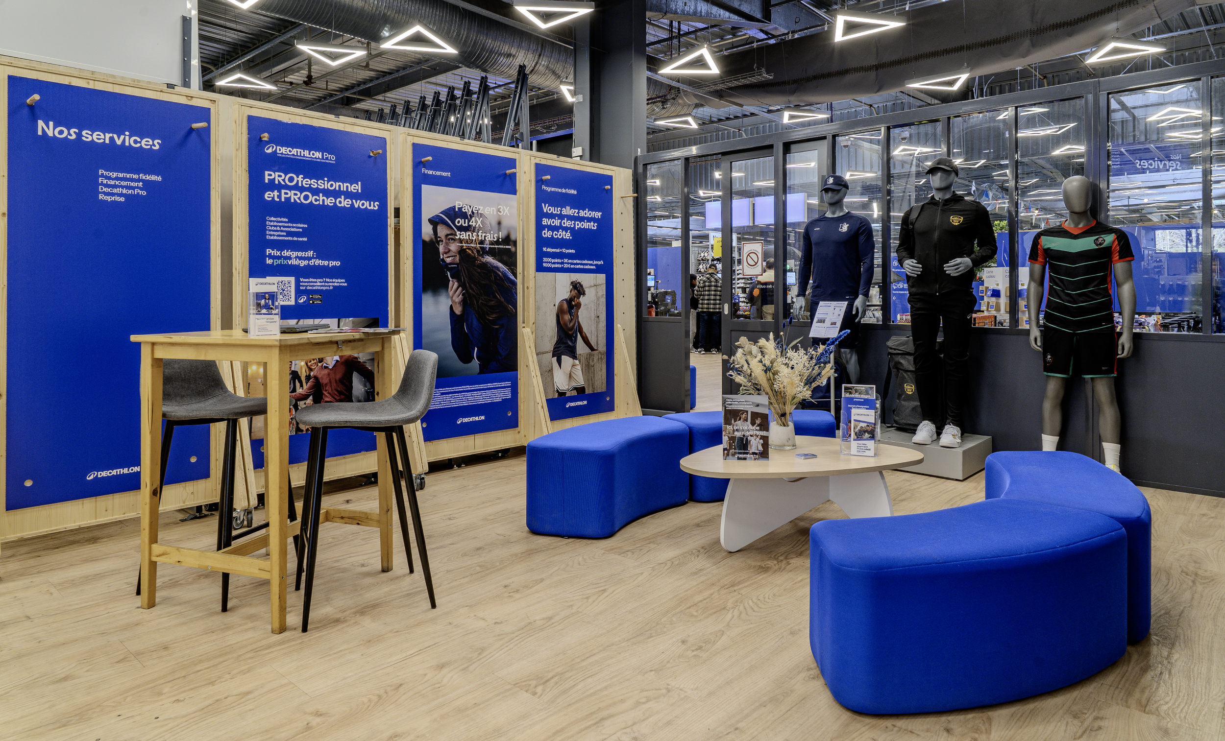 Espace d'accueil d'un magasin Decathlon avec des posters bleus, deux mannequins habillés en vêtements de sport, un coin salon avec des canapés bleus et une table, et une zone commerçante visible à l'arrière. Par Alexane Azé Architecture.