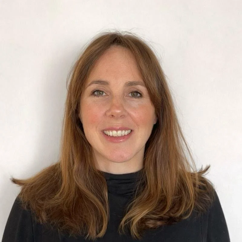 A woman with long, wavy reddish-brown hair, smiling, wearing a black top, standing in front of a plain white wall.