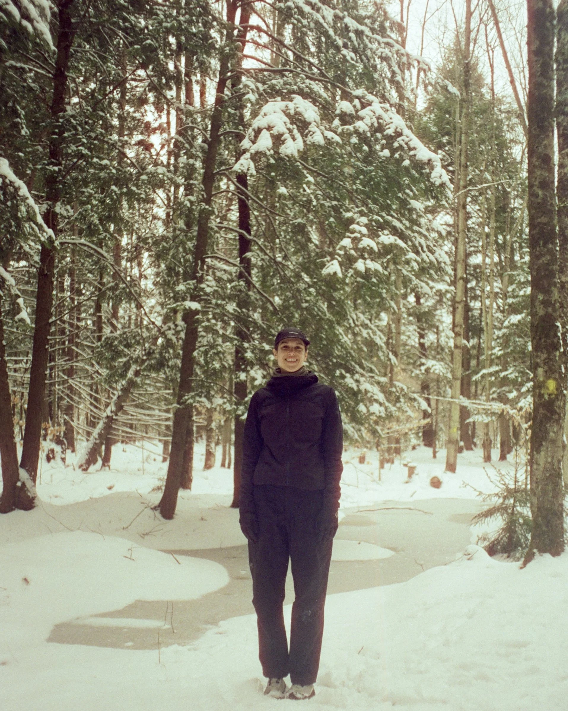 Une femme souriante se tenant dans une forêt enneigée avec des arbres couverts de neige.