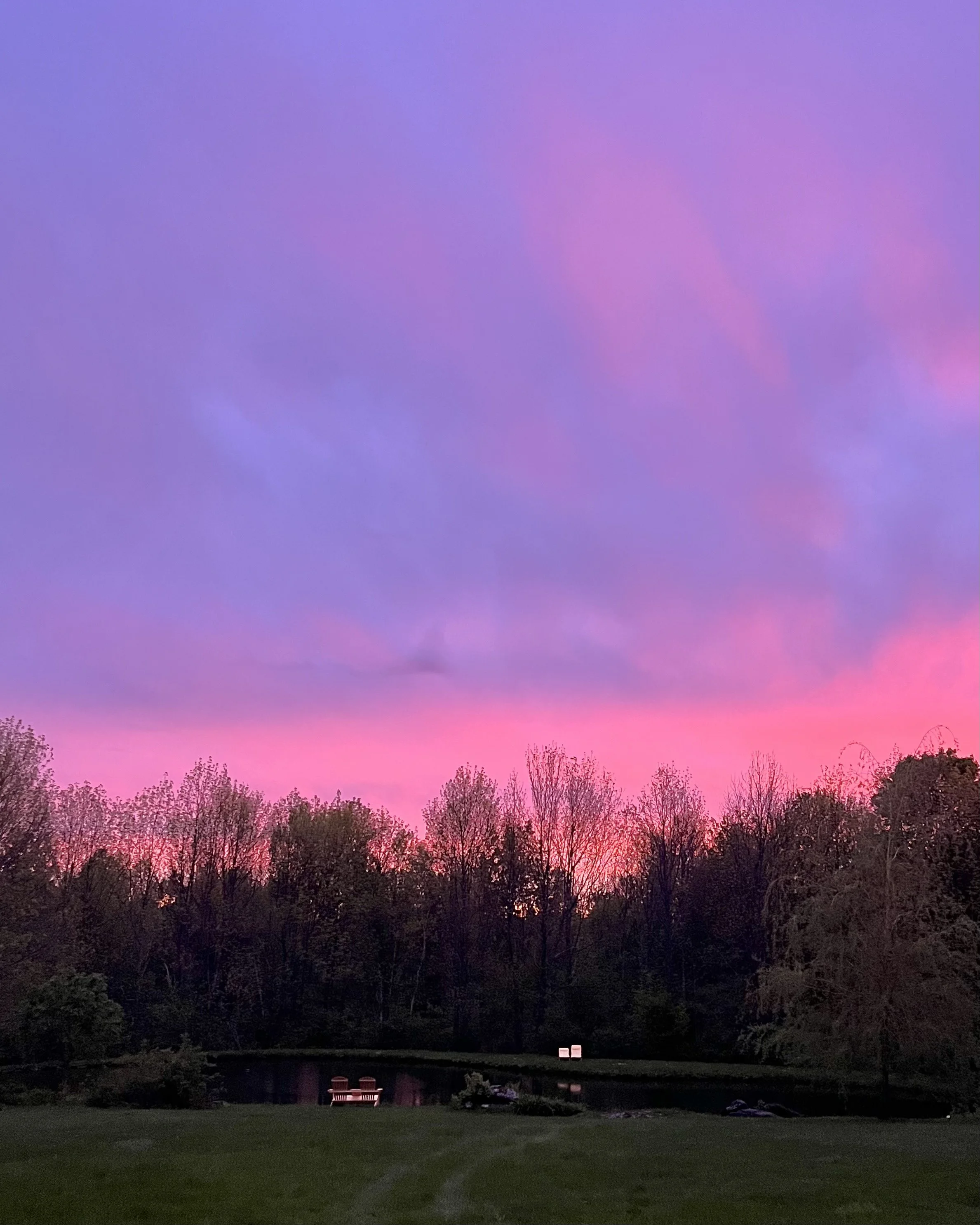 Ciel coloré au coucher du soleil avec des teintes violettes, roses et bleues, silhouettes d'arbres nus, un étang réfléchissant et deux chaises près de l'eau.