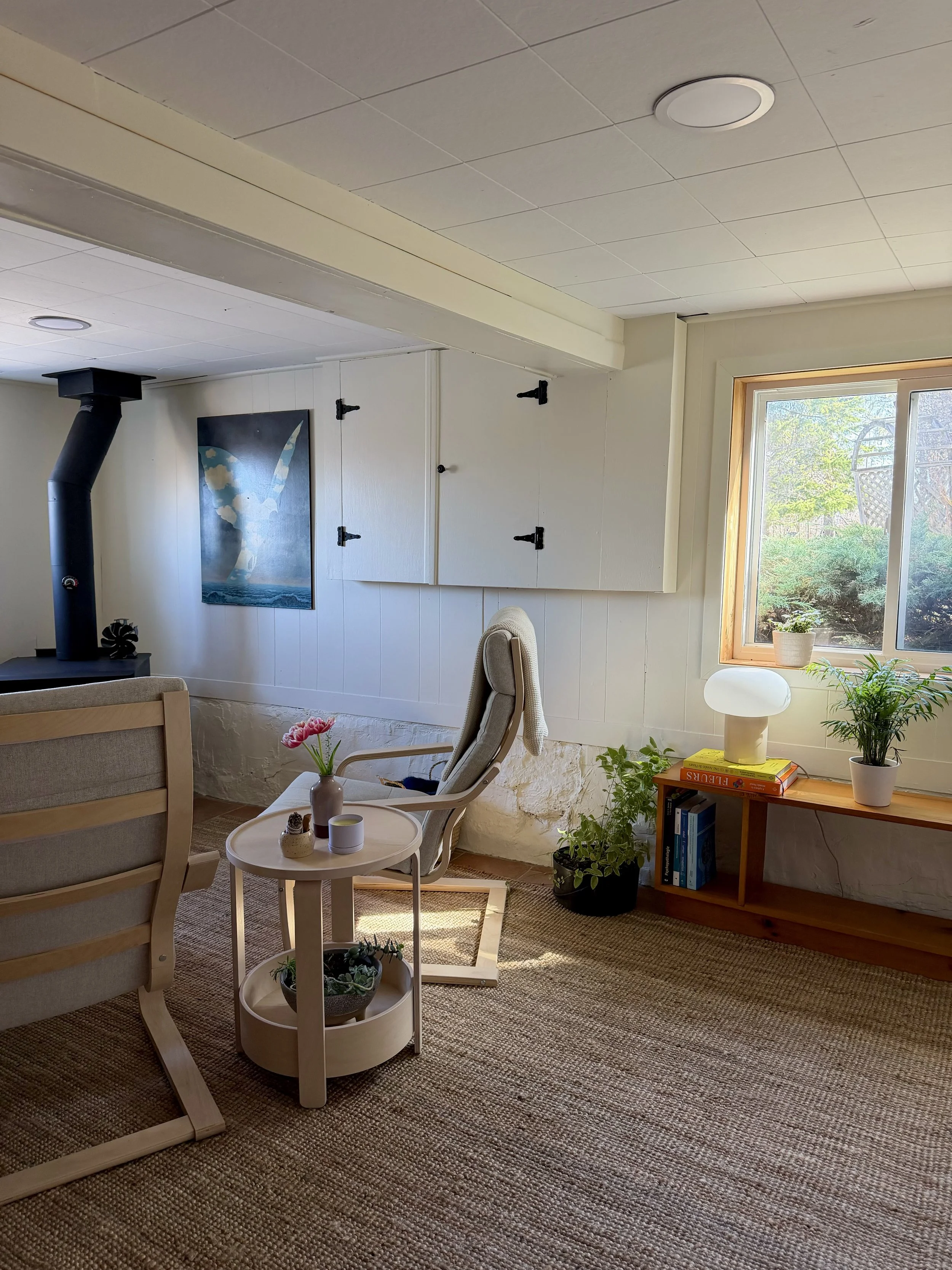 Intérieur d'une pièce lumineuse avec fenêtre laissant entrer la lumière naturelle, décorée avec des plantes et des meubles en bois, tableau d'oiseau sur le mur, tapis beige et fauteuils en bois avec coussins, un petit meuble avec livres et une lampe