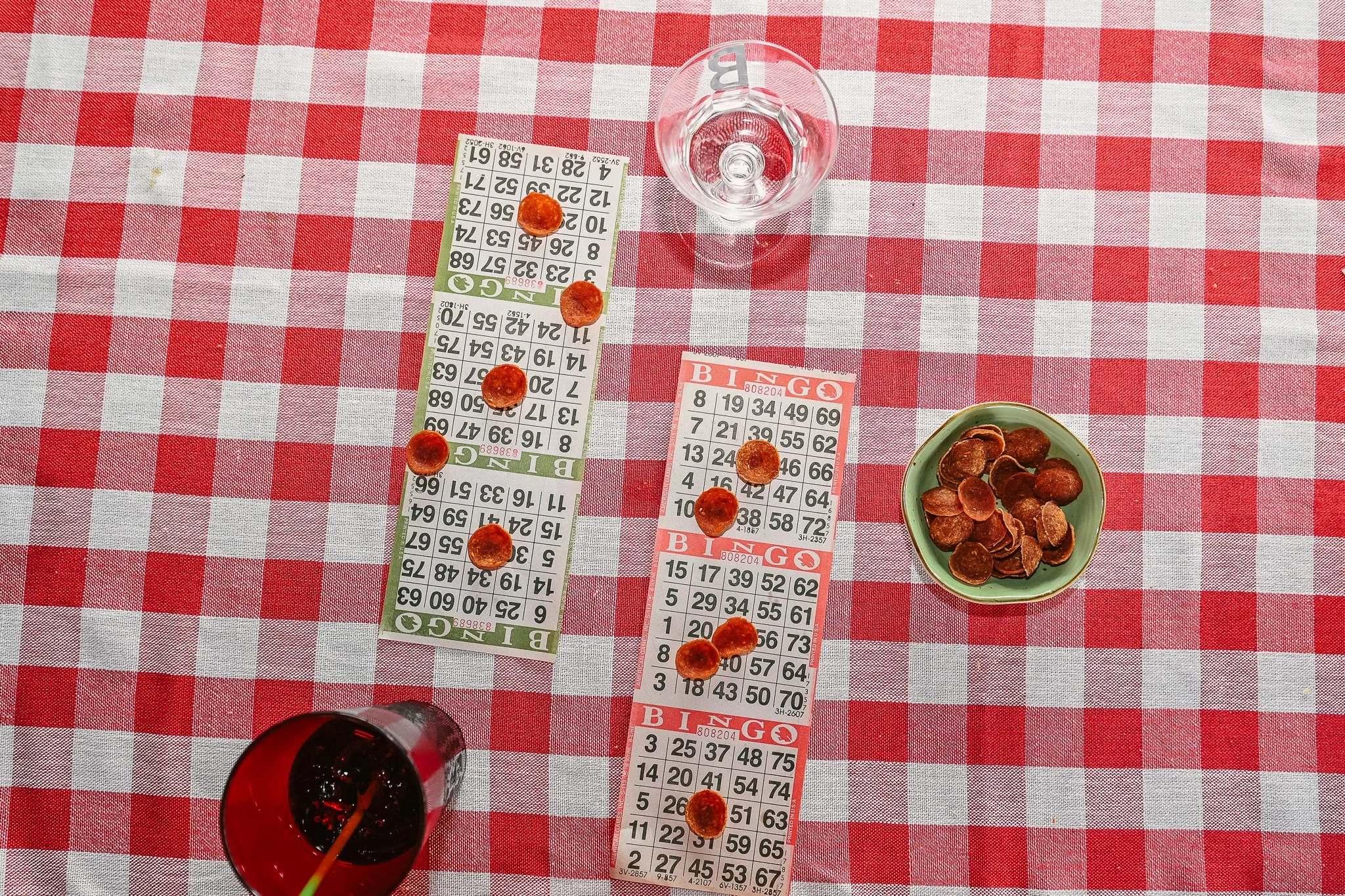 Bingo cards, pepperoni slices, a glass of water, and a drink on a red and white checkered tablecloth.