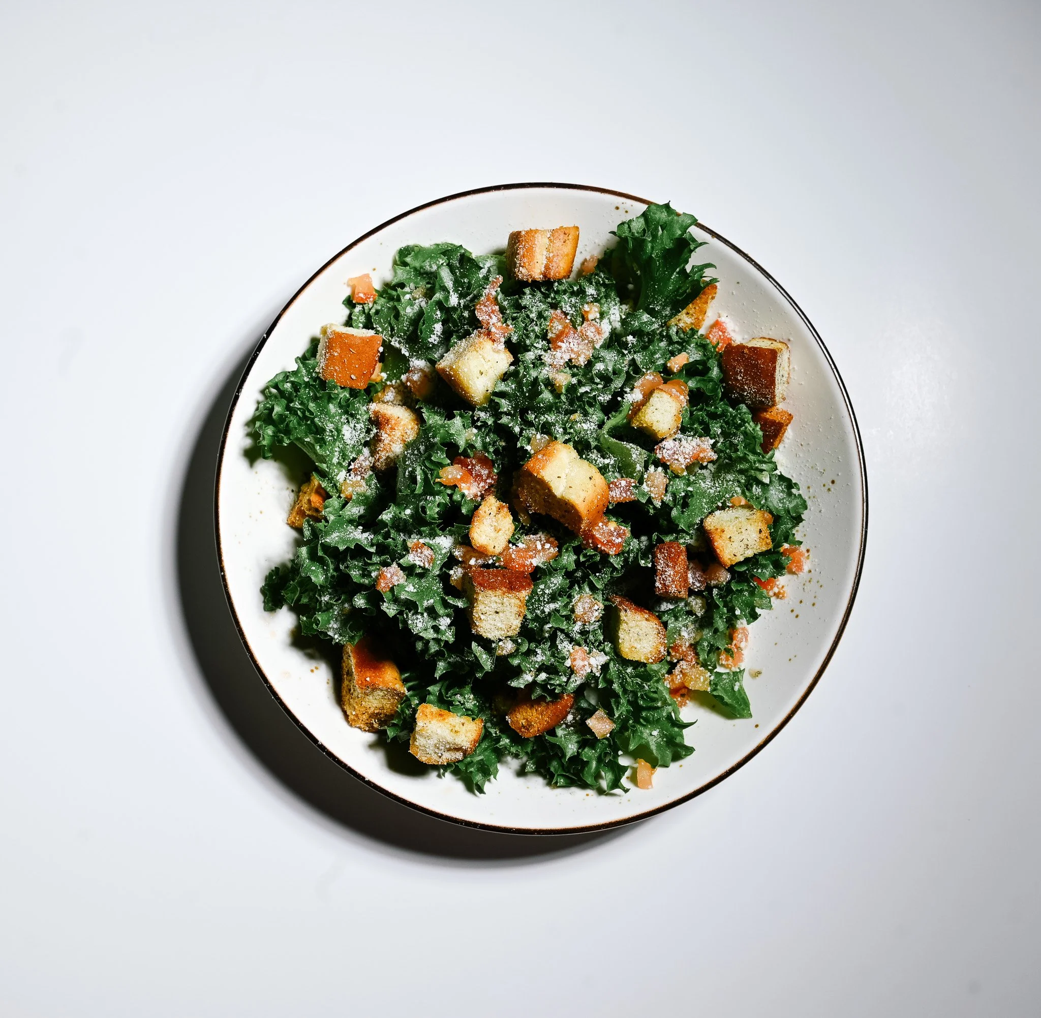 Kale Caesar salad with croutons and grated cheese in a white bowl on white background.