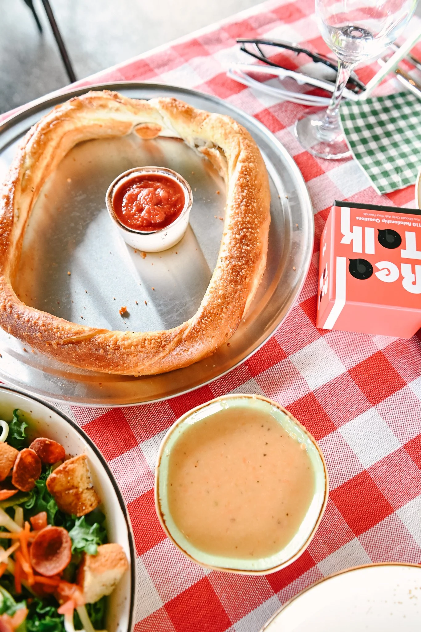 O-shaped bread with a small container of marinara sauce in the center, on a metal tray, with a bowl of salad, a cup of soup, a wine glass, and a red and white checkered tablecloth.