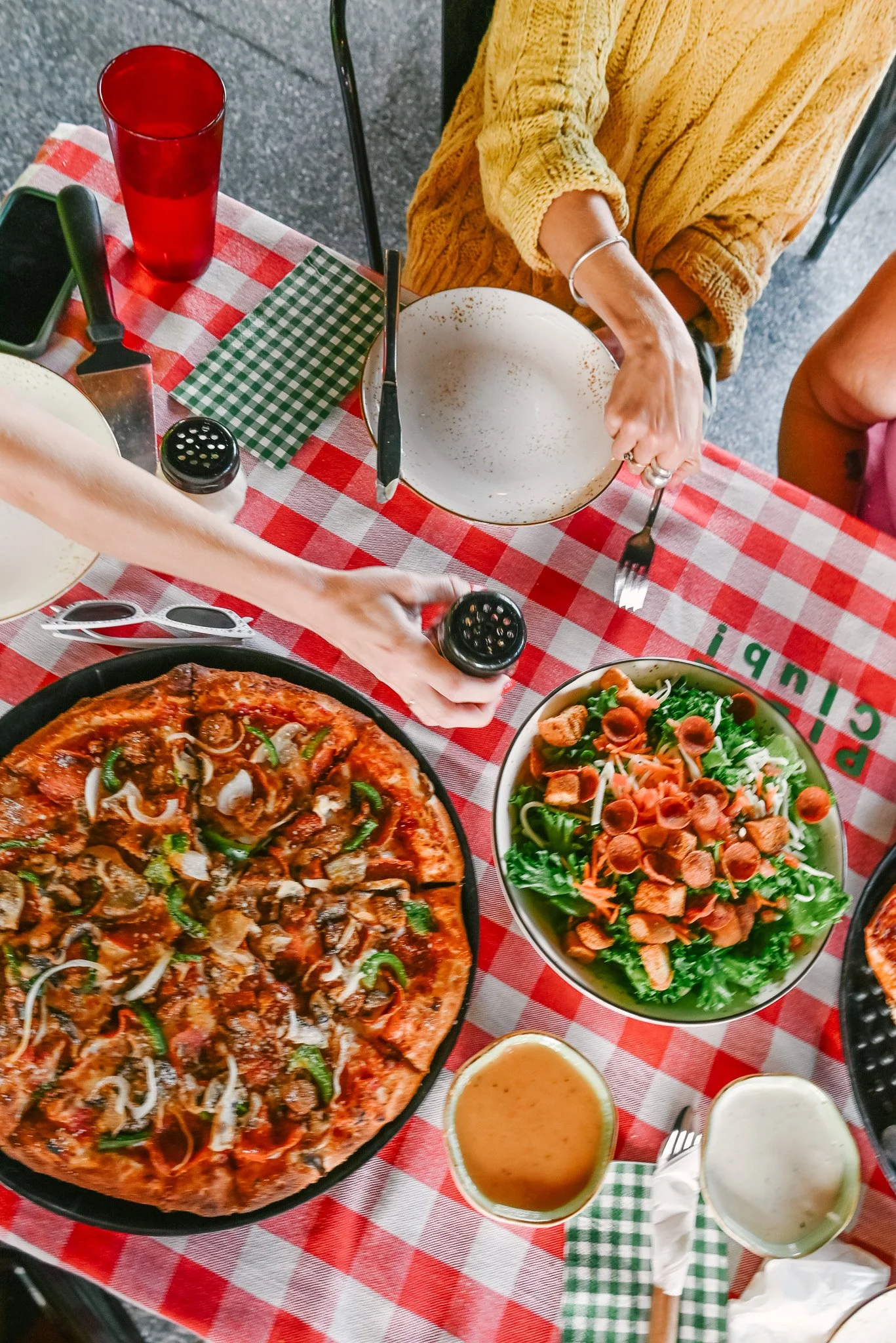 A top-down view of a table with a large pizza, a bowl of salad with lettuce, cherry tomatoes, and carrots, two small bowls of dressing, a pair of sunglasses, and a red drinking glass. Two people are reaching for items on the table, with one wearing a yellow sweater.