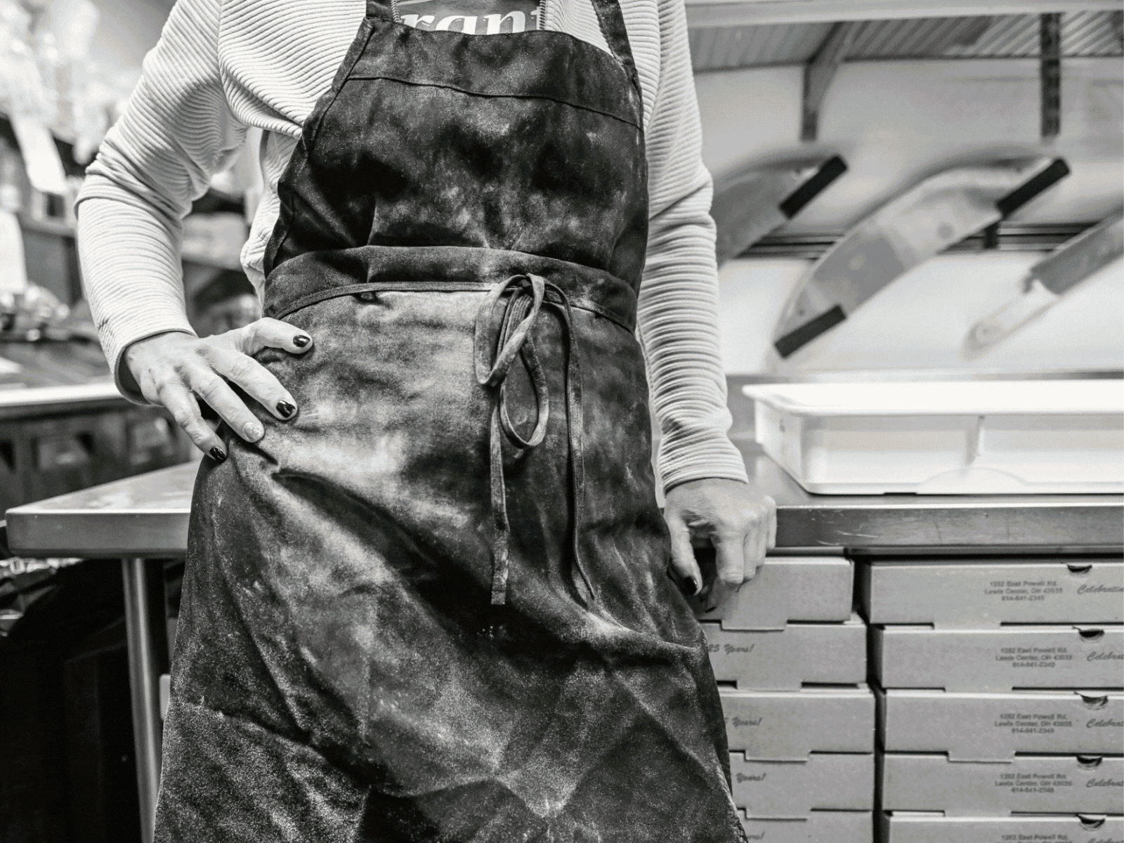 Person wearing an apron standing in a kitchen or bakery, with stacked boxes and trays of baked goods or food items in the background.