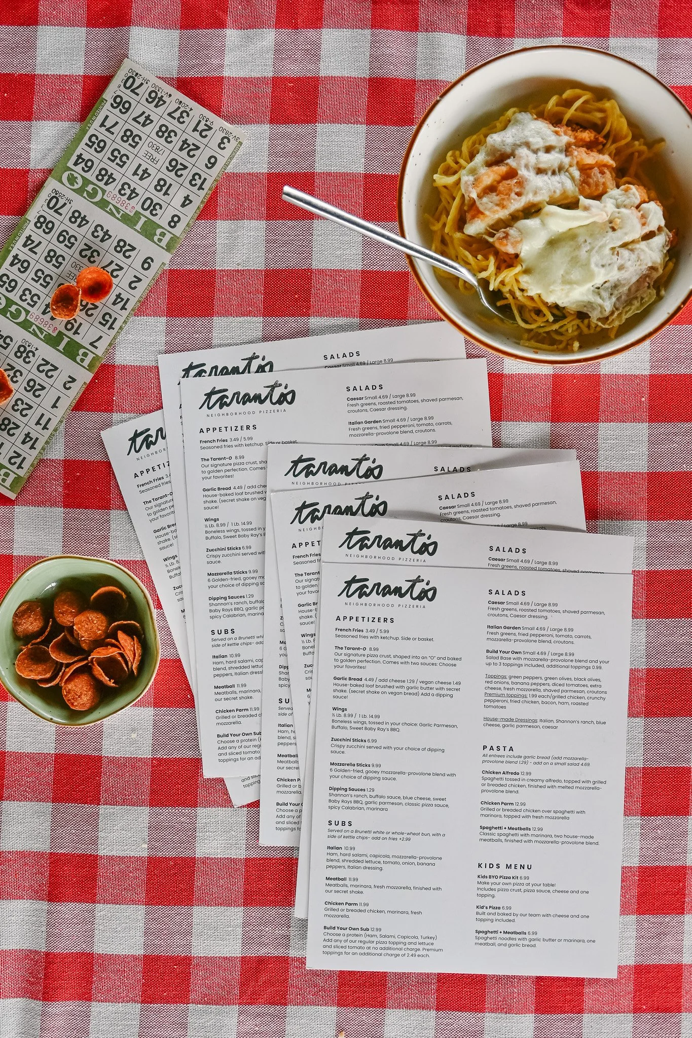 A table setting at a pizzeria with a bowl of spaghetti topped with sauce and cheese, a small bowl of pepperoni slices, multiple printed menus, a bingo card, and the checkers are all on a red and gray checkered tablecloth.
