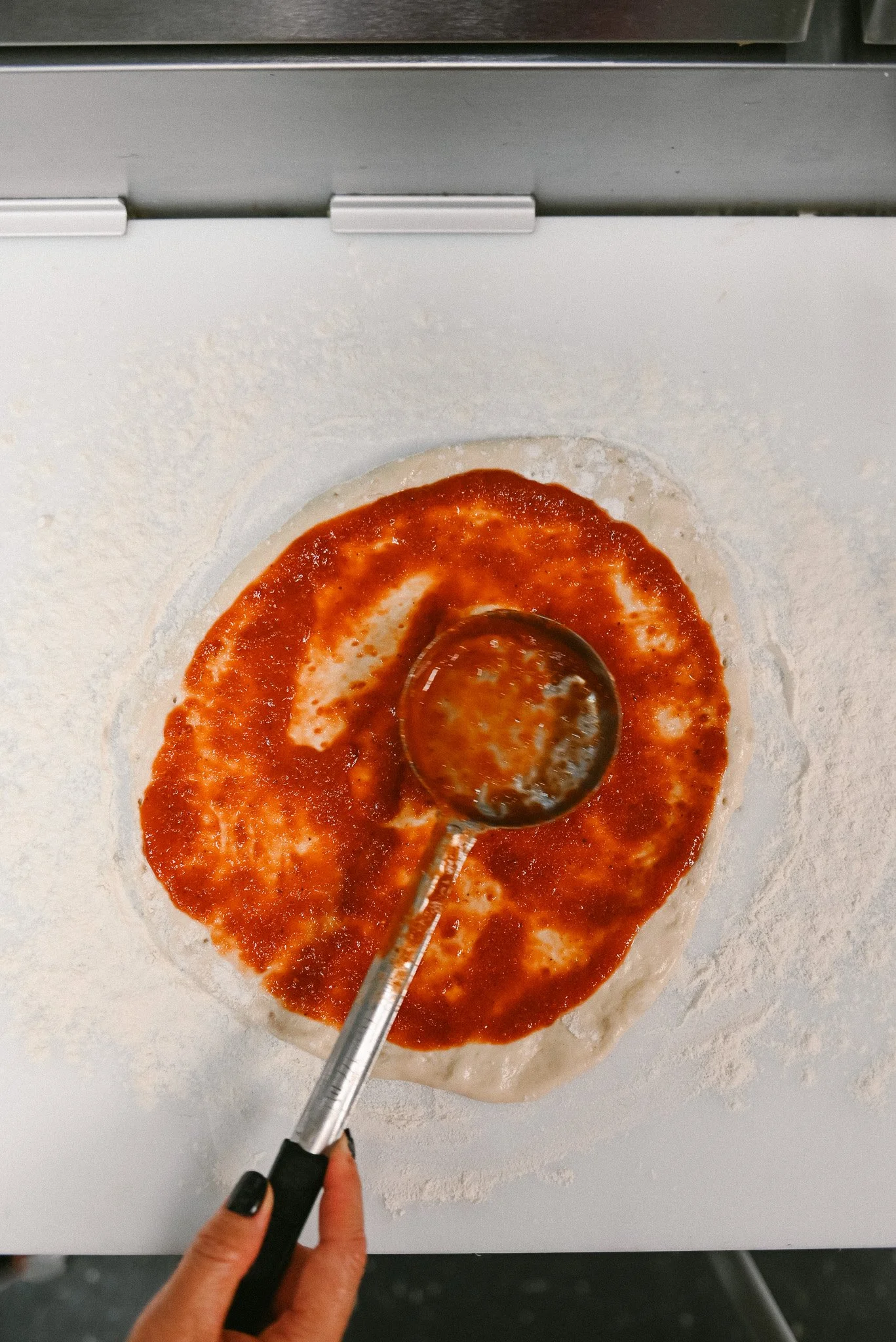 A person spreading marinara sauce onto pizza dough at a kitchen table.