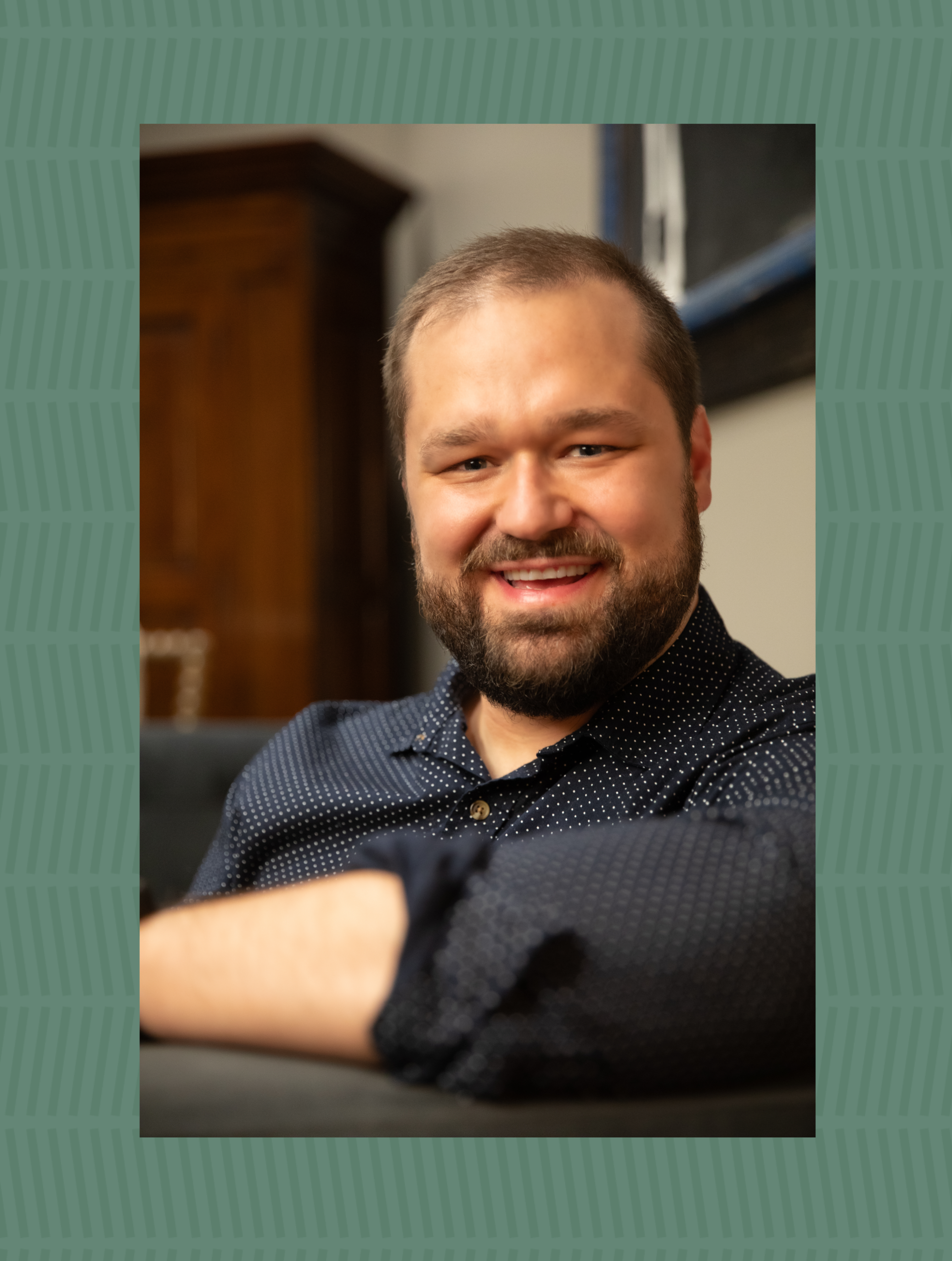 A smiling man with a beard and short hair, wearing a dark dotted shirt, posed indoors with a blurred background.