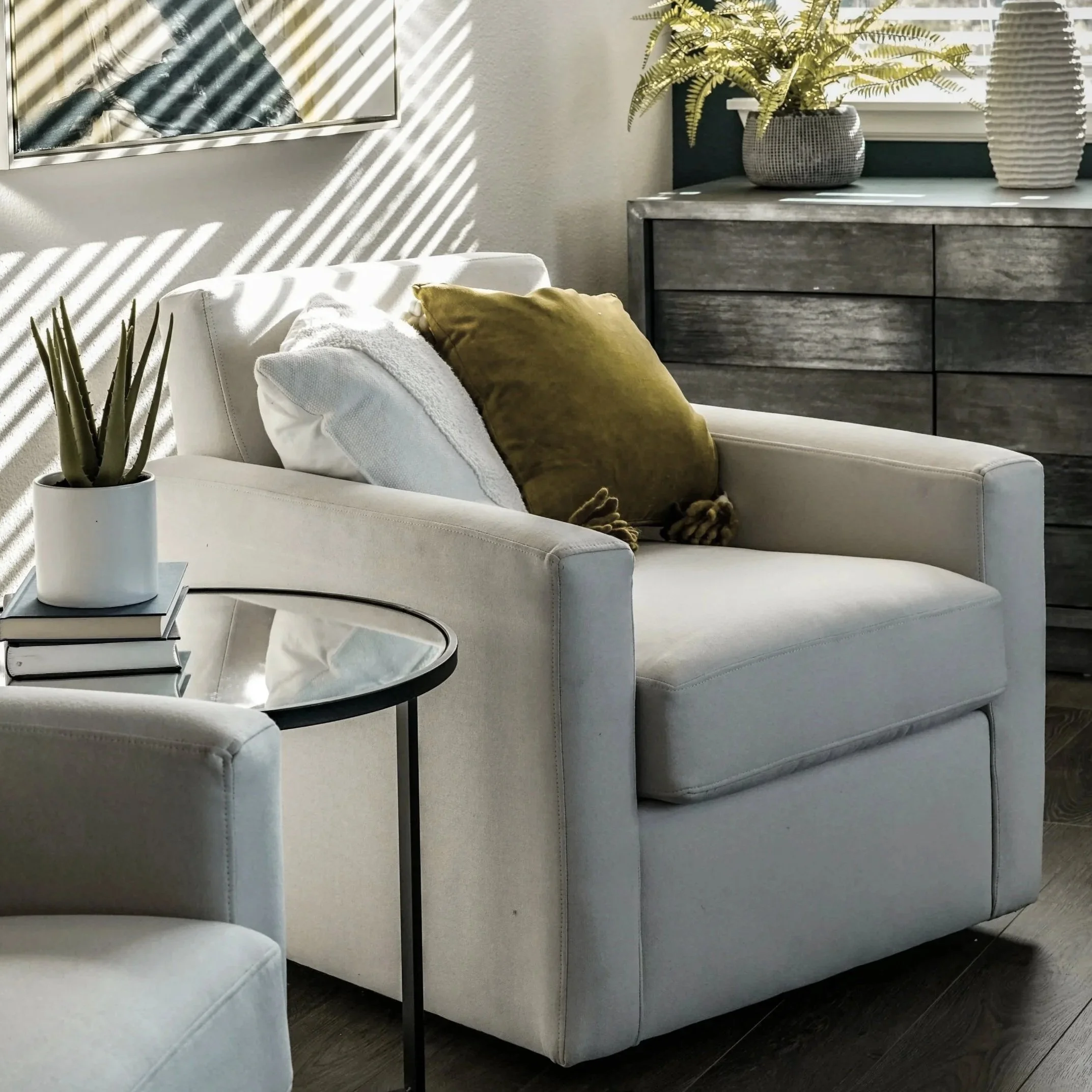 A cozy living room corner with a white armchair, decorated with a green pillow and a white blanket, next to a small round black table with a potted aloe vera and a stack of books. Sunlight creates striped shadows on the wall.