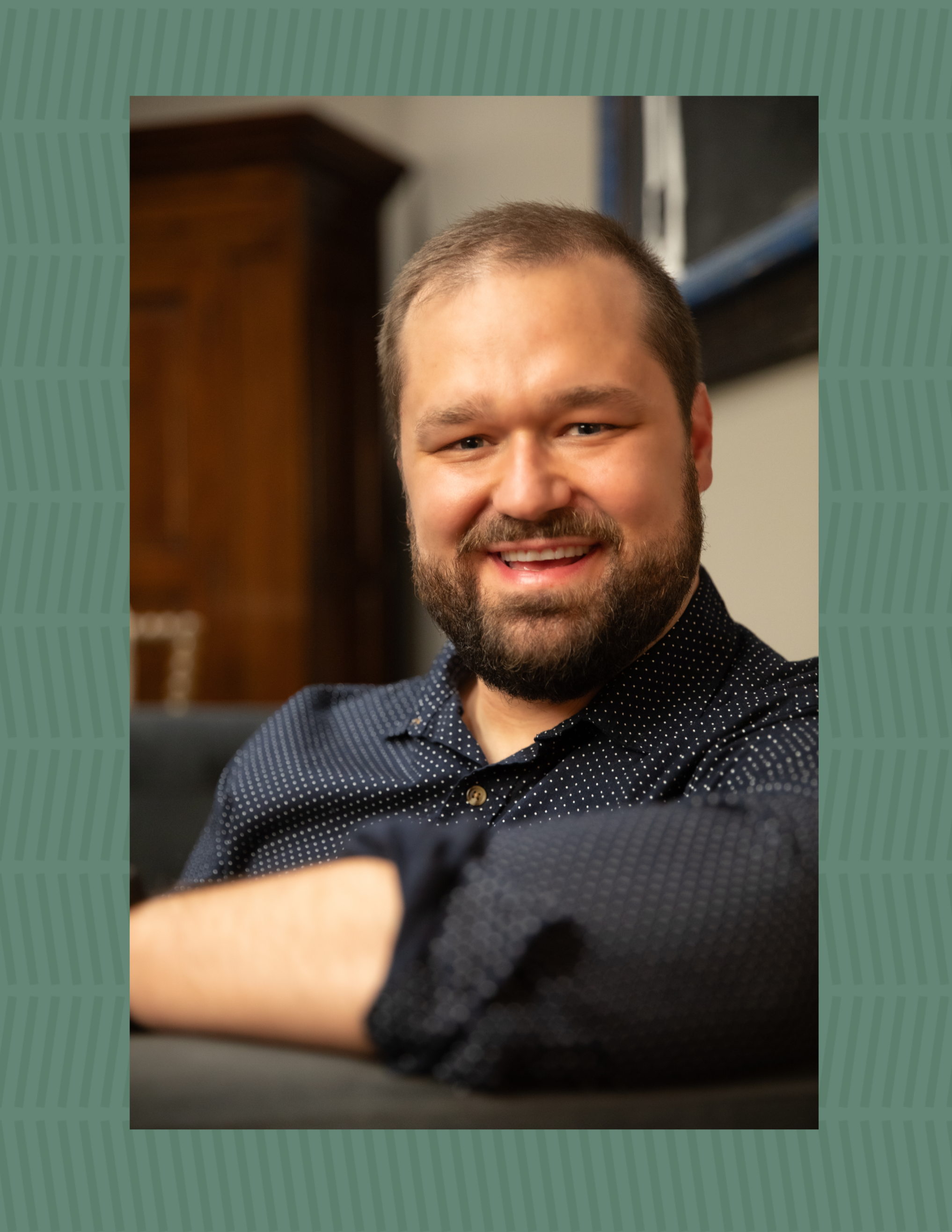A smiling man with a beard and short hair wearing a black, dotted button-up shirt, sitting indoors with a blurred background of a wooden cabinet and a white wall.