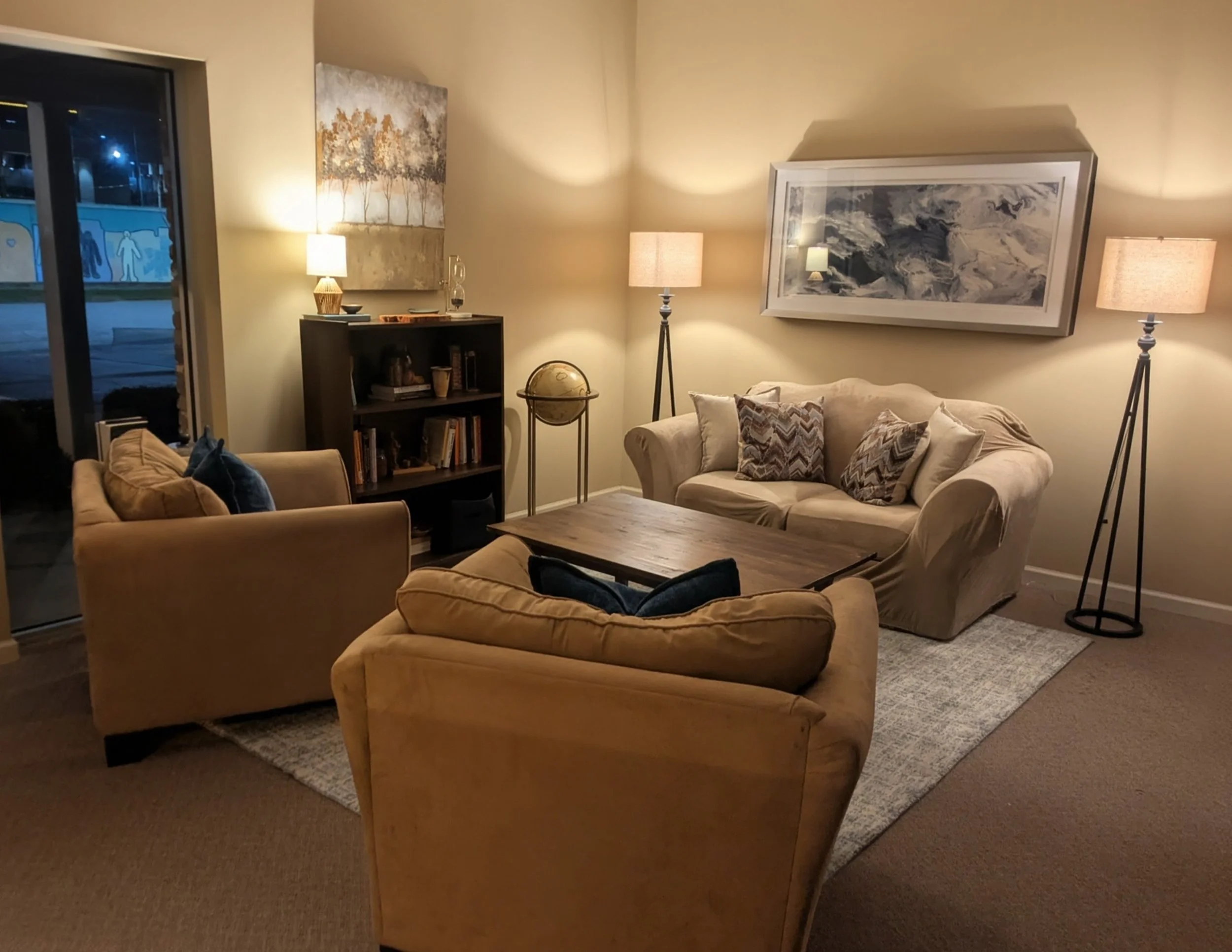 Living room with beige sofas, a wooden coffee table, a black bookshelf, and two tall lamps. Artwork hangs on the walls, one abstract and one landscape. Glass sliding door to the outside, night time.