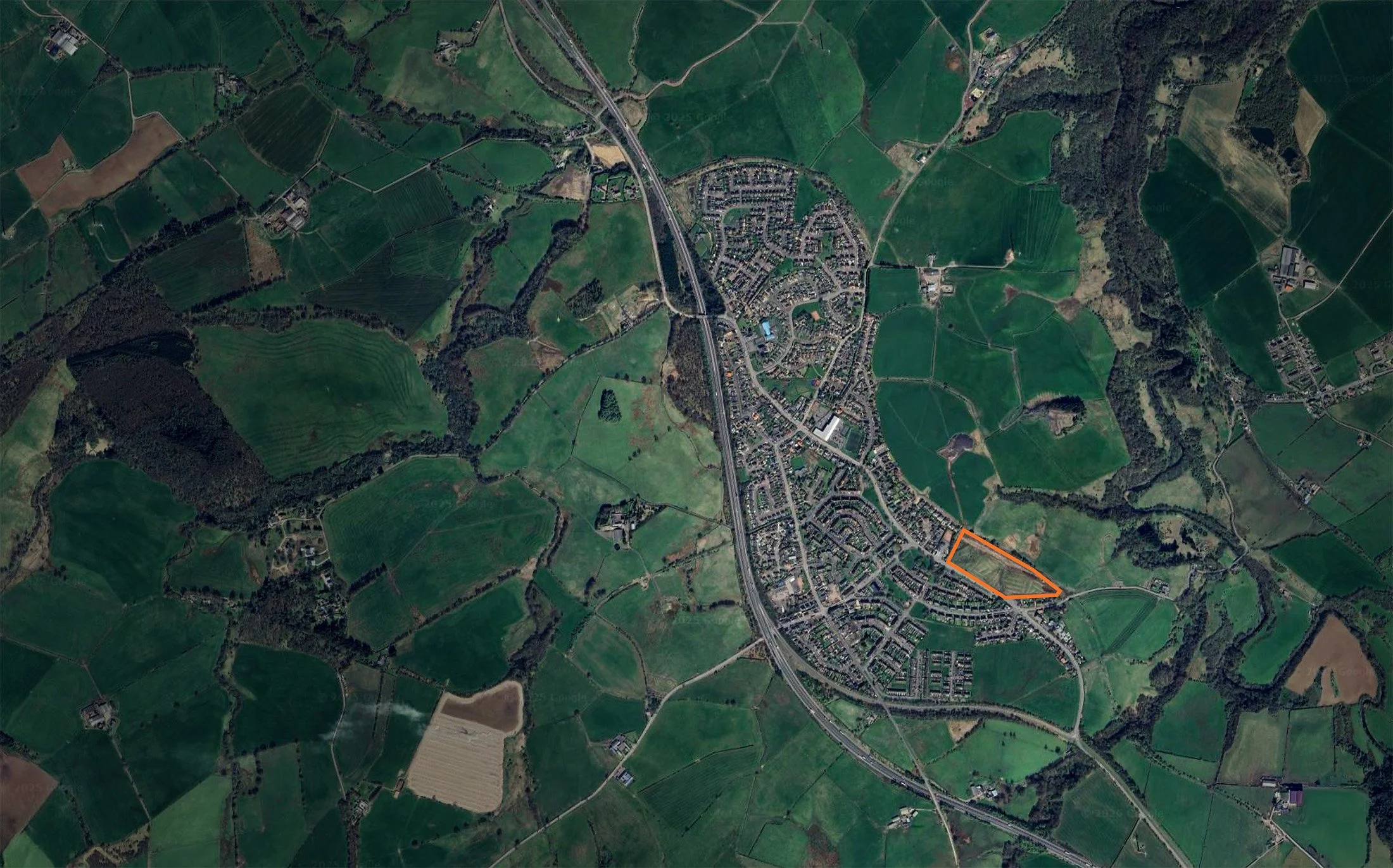 Satellite view of a town with surrounding green fields and roads, with an outlined triangular plot of land in the southeastern part of the town.